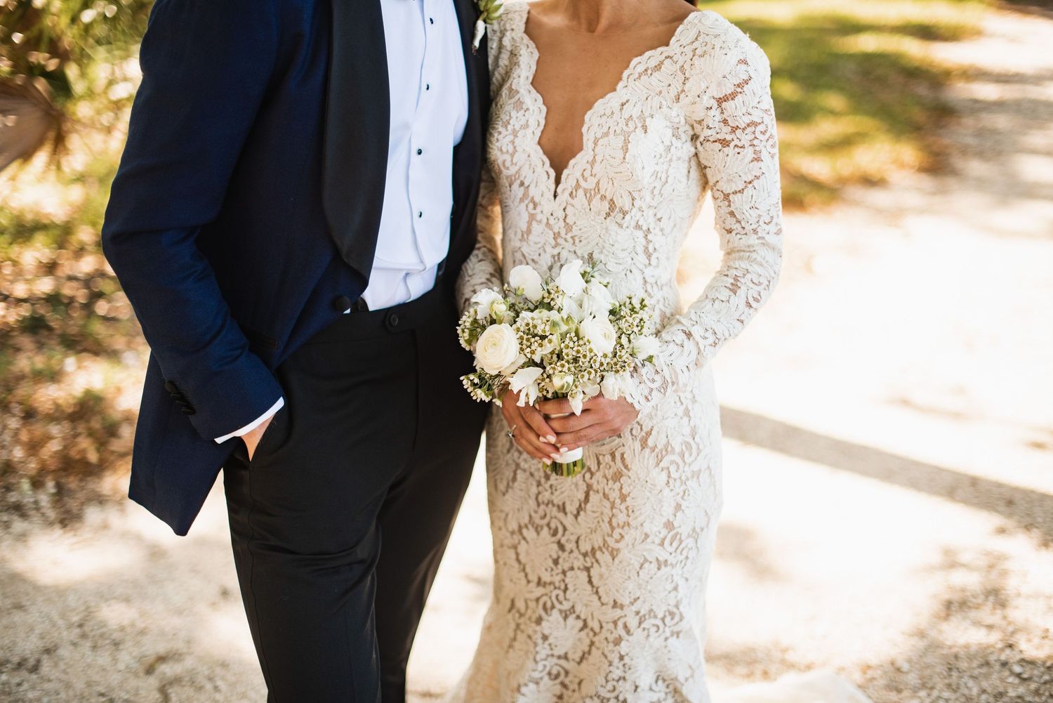 A couple in wedding attire stands together in a sun-dappled outdoor setting, the bride wearing an elegant lace gown.