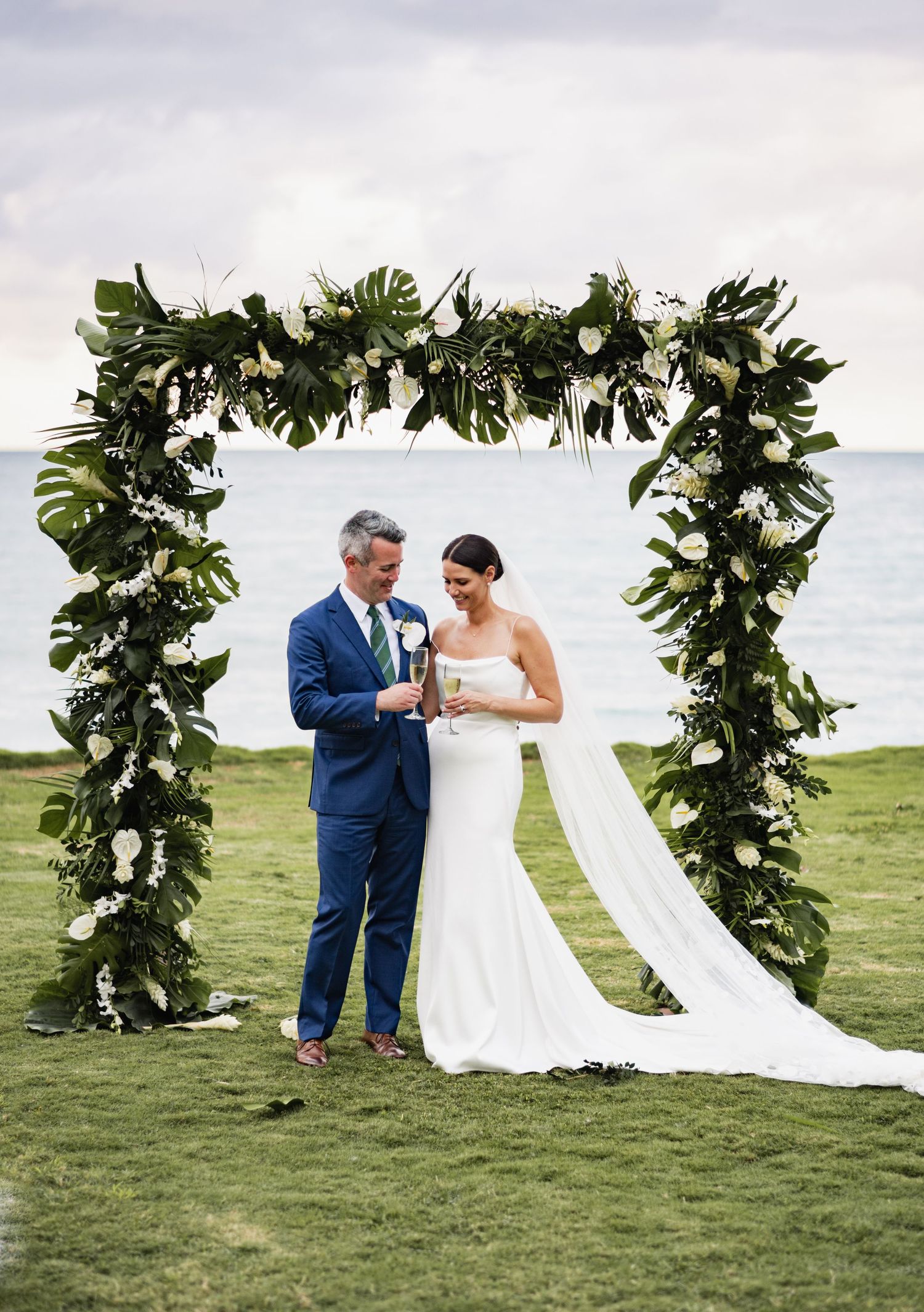 A wedding ceremony takes place on a grassy oceanfront with a lush green floral arch adorned with tropical leaves and flowers.