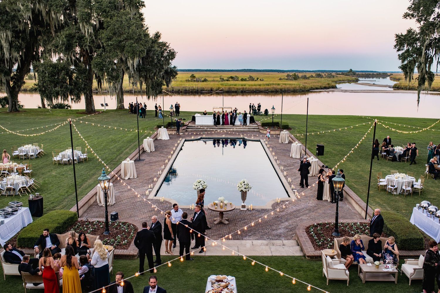 Outdoor wedding ceremony setup with white chairs on a green lawn overlooking a scenic waterfront at sunset.