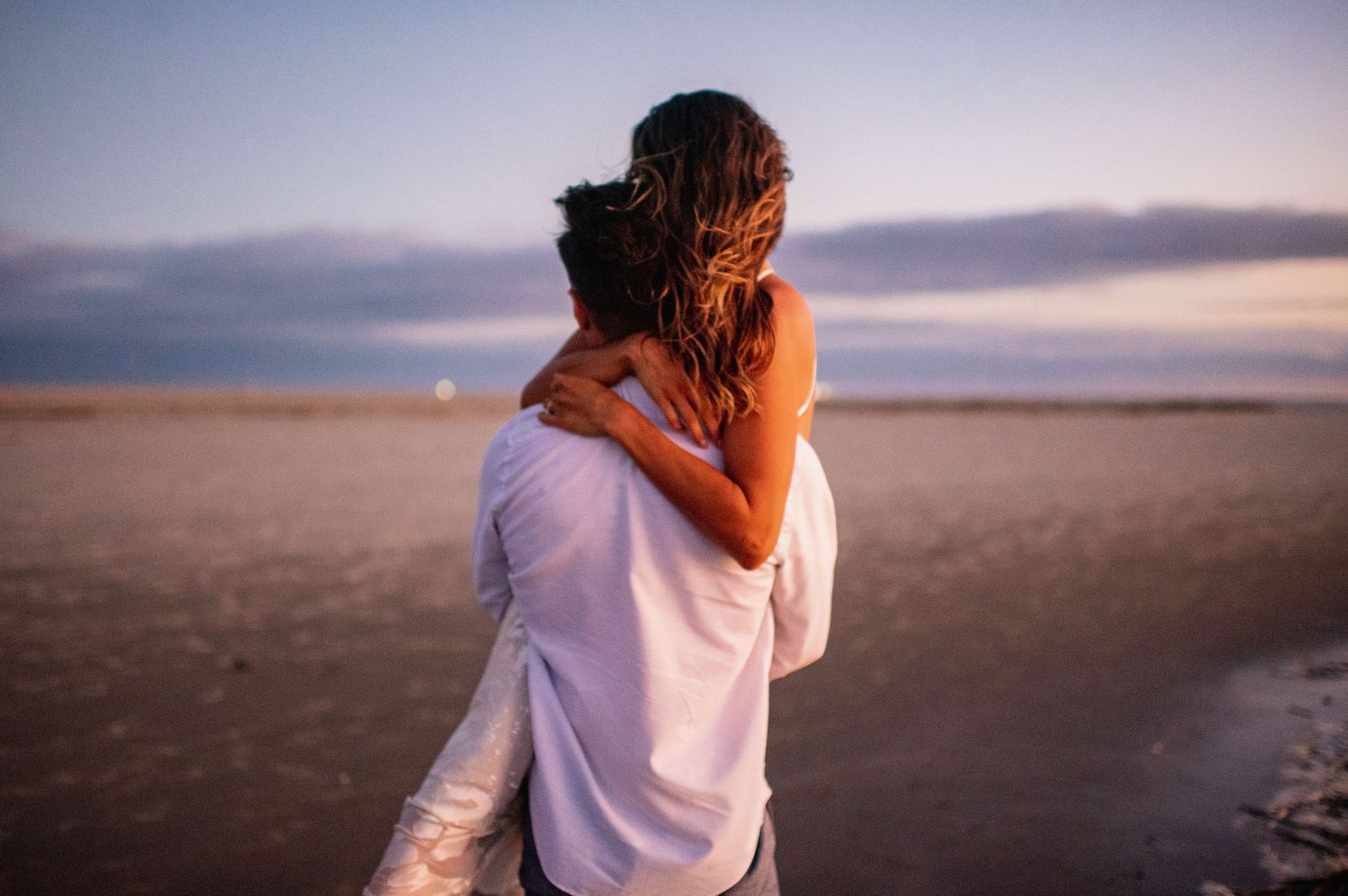 A couple embraces on an empty beach at sunset with warm golden light illuminating the scene.