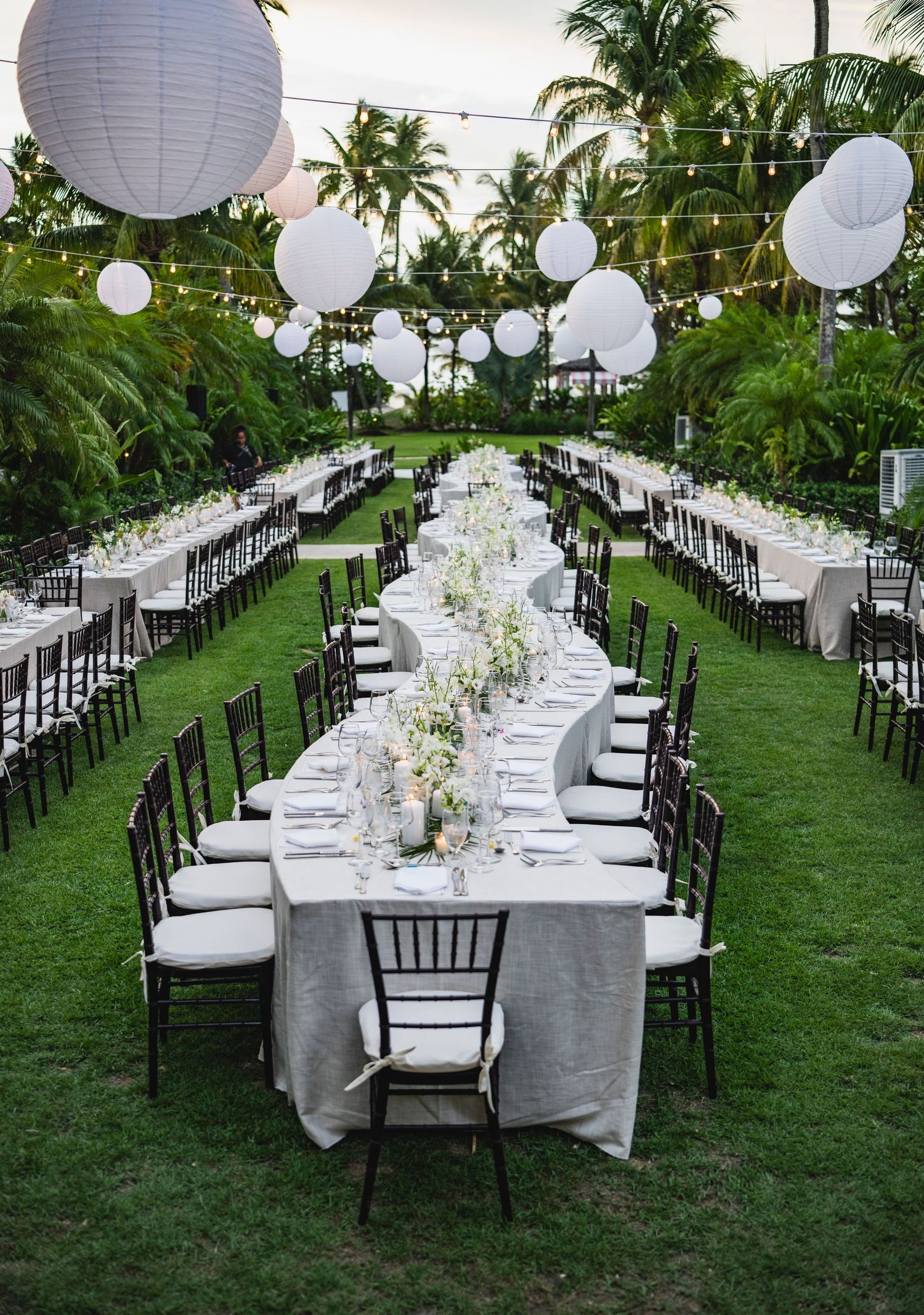 Elegant outdoor wedding reception setup with long tables, white decor, paper lanterns and palm trees in a tropical garden setting.