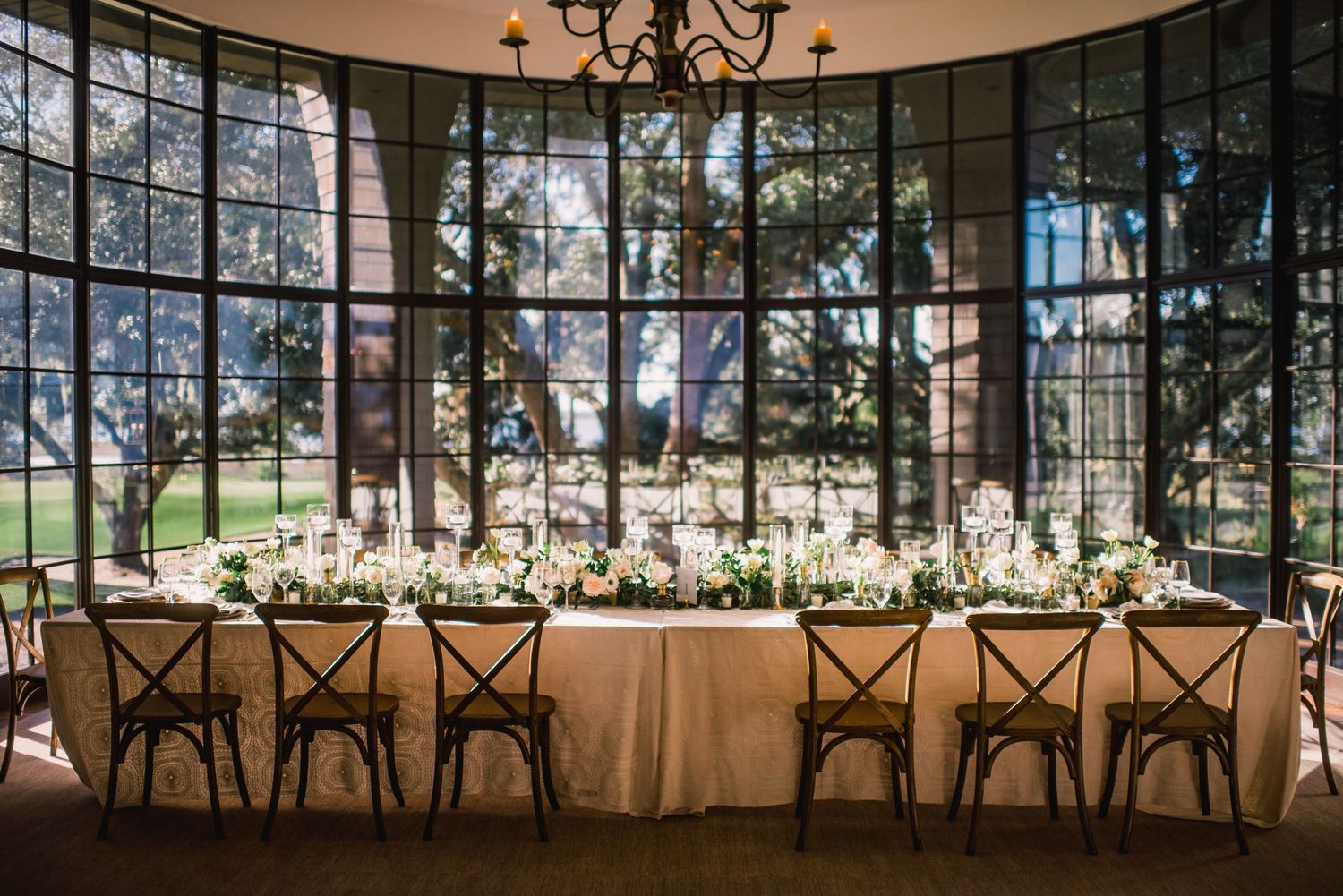 Elegant wedding reception setup in a glass-walled venue with long tables and white floral centerpieces under chandeliers.