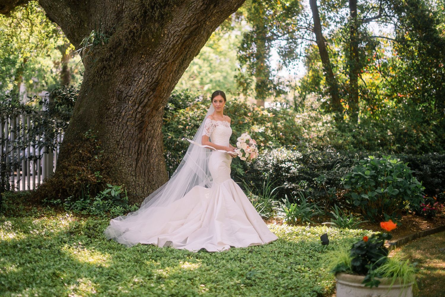 A figure in a pink ballgown poses gracefully in a lush garden setting beneath towering trees and dappled sunlight.