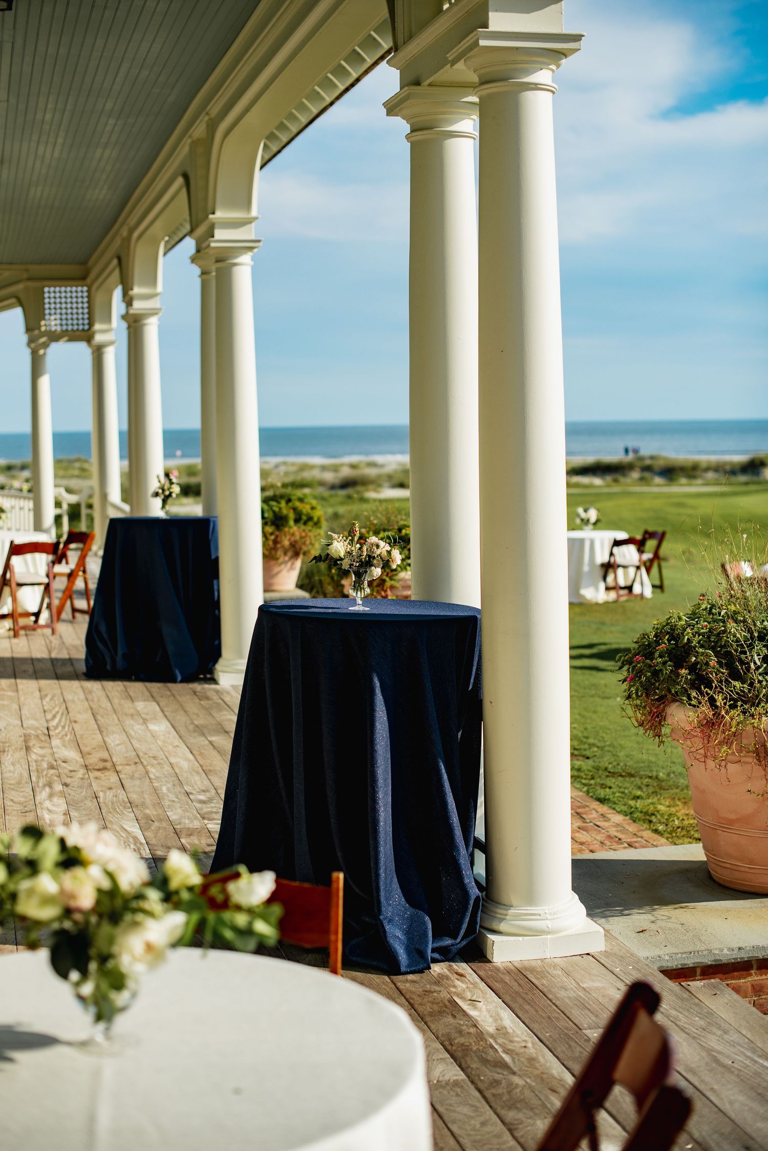 A covered veranda with white columns and navy blue tablecloths overlooks a scenic coastal golf course on a sunny day.