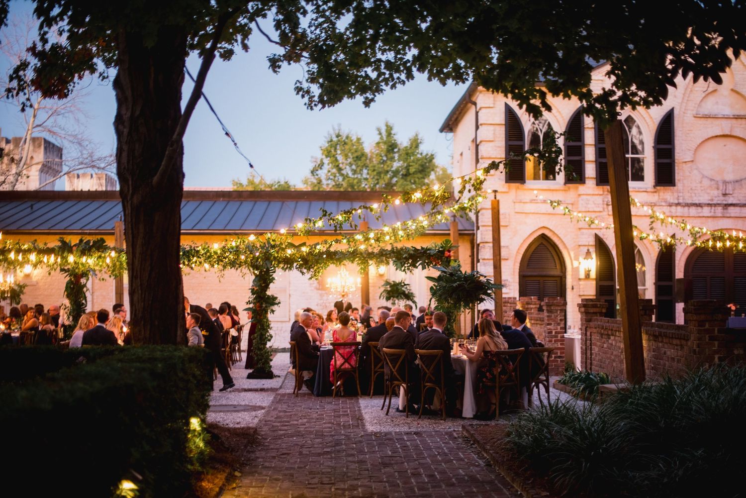 String lights illuminate a rustic stone courtyard with arched doorways and windows during an evening event.