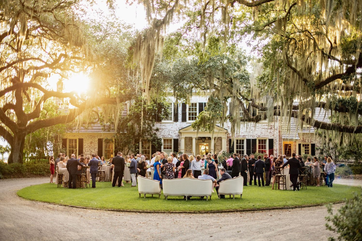 Wedding guests gather on a scenic plantation property at sunset with Spanish moss draped oak trees and a historic home.