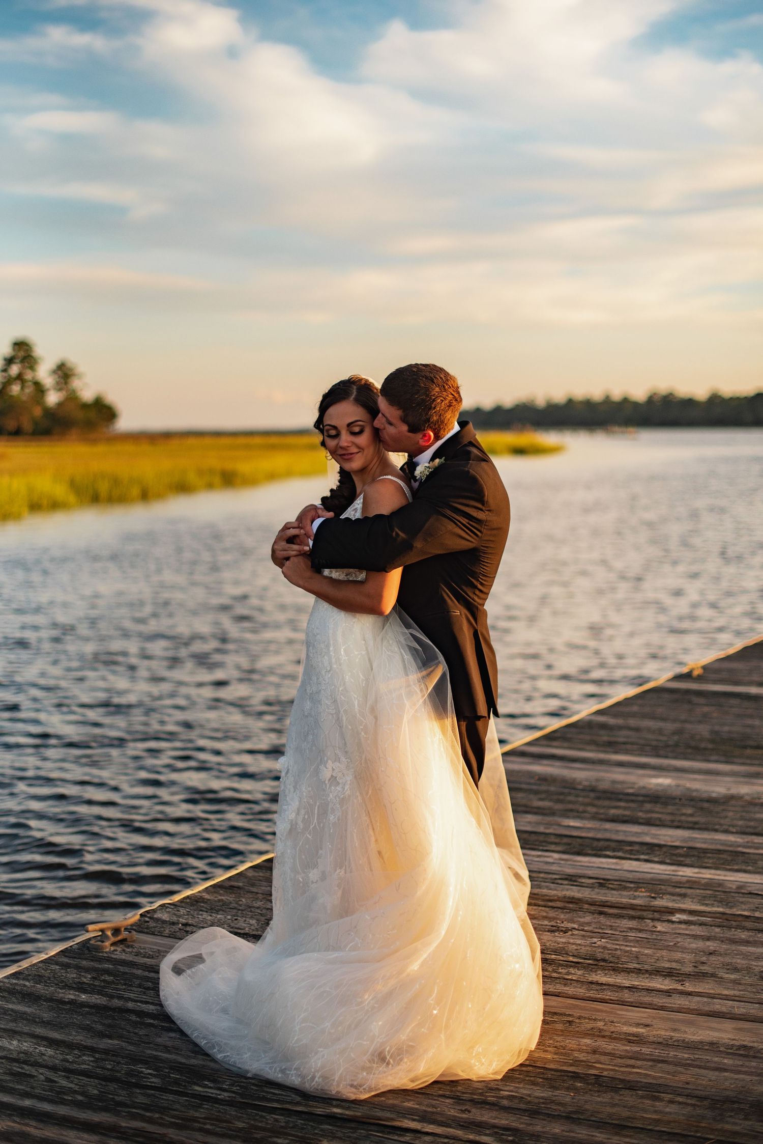 A couple shares an intimate moment at sunset on a wooden dock overlooking a serene waterfront.