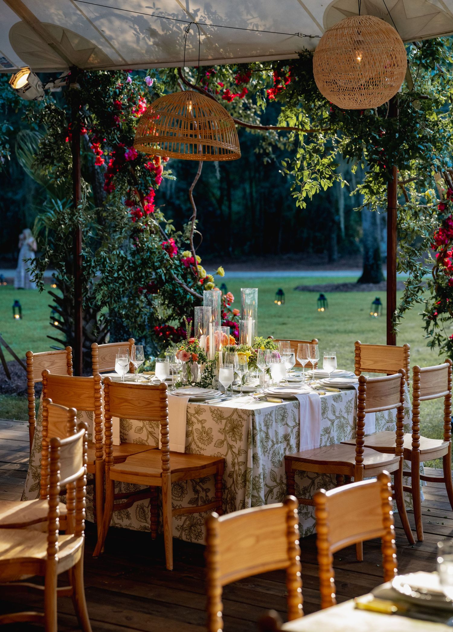 Elegant outdoor dining setup with wooden chairs and woven pendant lights under a natural canopy of trees and flowers.