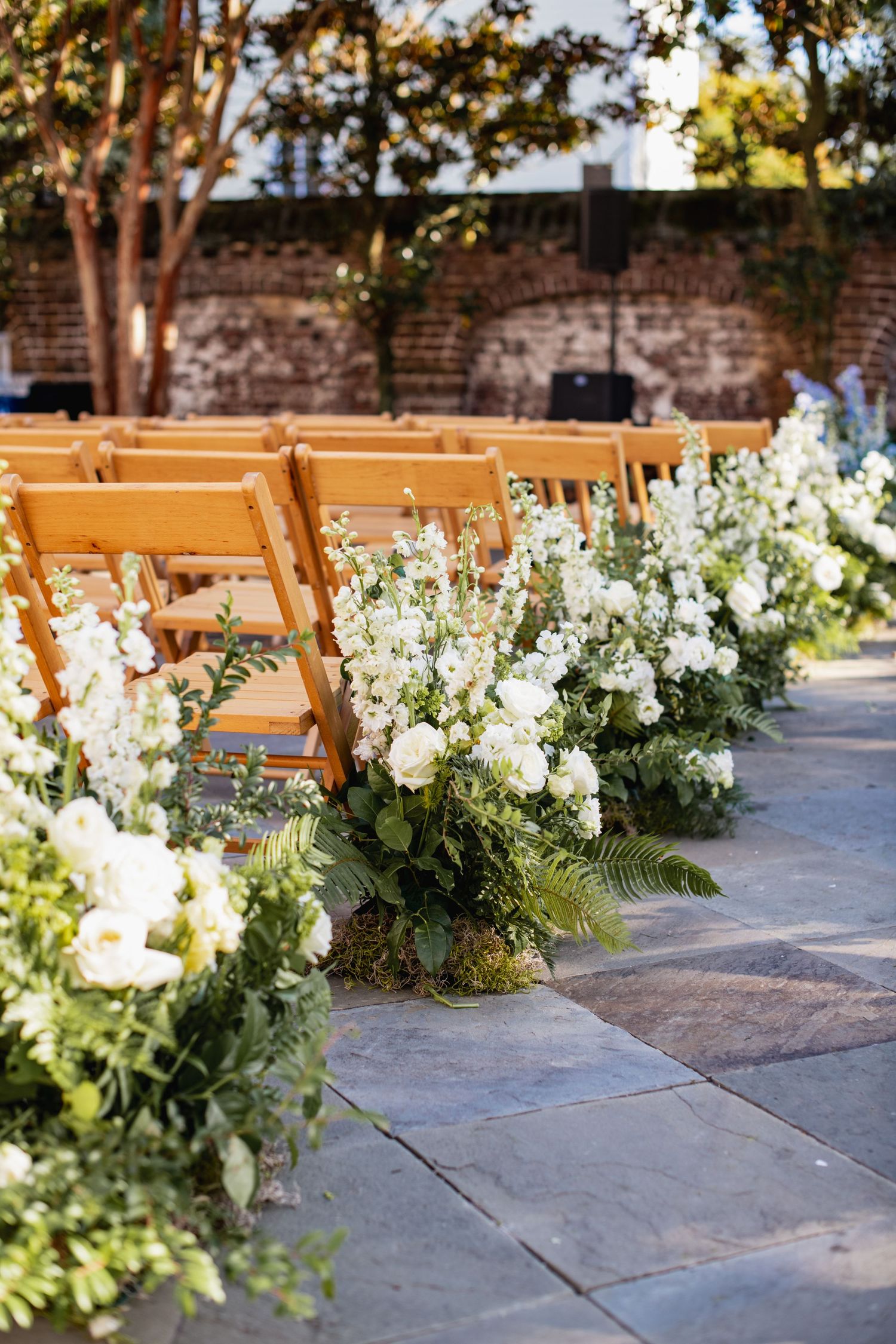 Wooden folding chairs line an outdoor ceremony aisle decorated with lush white floral arrangements and greenery.