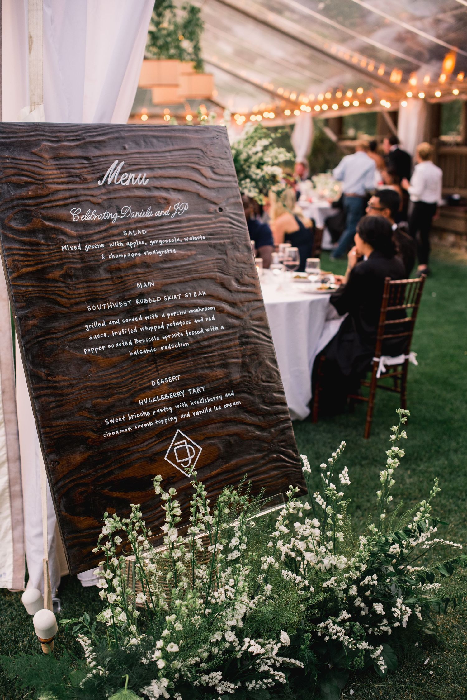Rustic wooden wedding menu signs displayed among delicate white flowers and string lights at an outdoor reception venue.