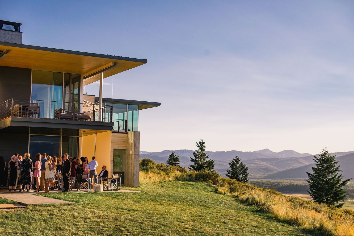 Modern hillside home with expansive windows overlooks a scenic mountain landscape at sunset.