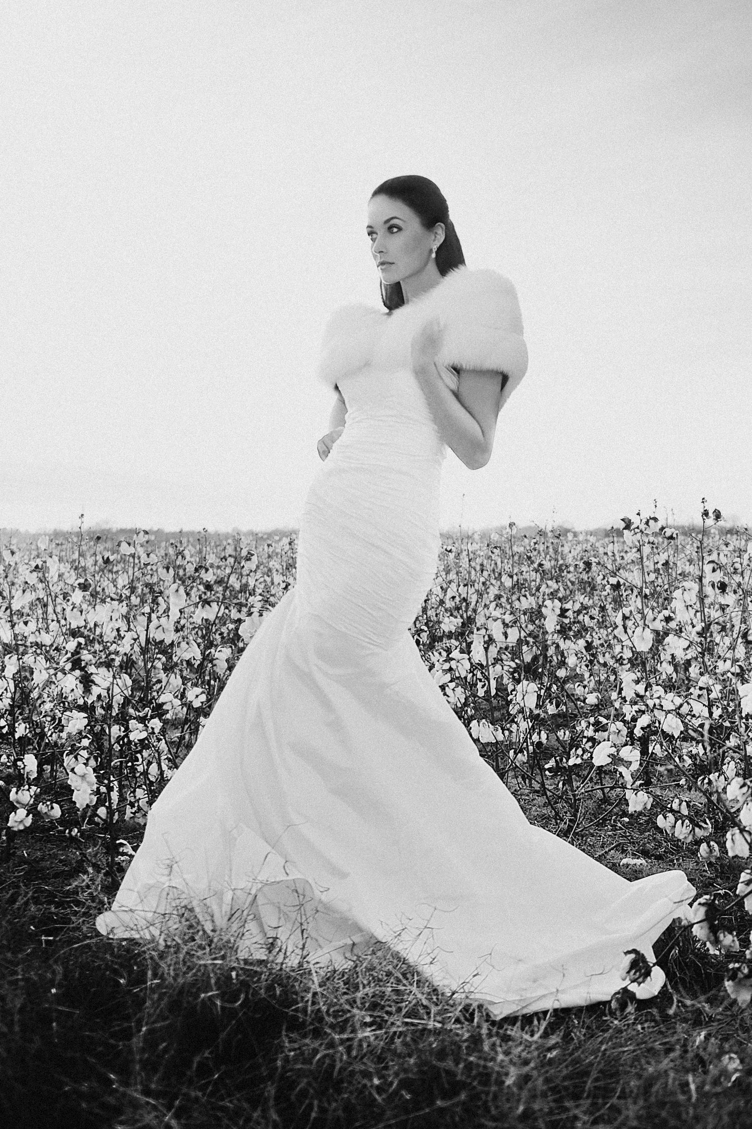 A bride in a flowing white dress twirls gracefully through a blooming cotton field in black and white photography.