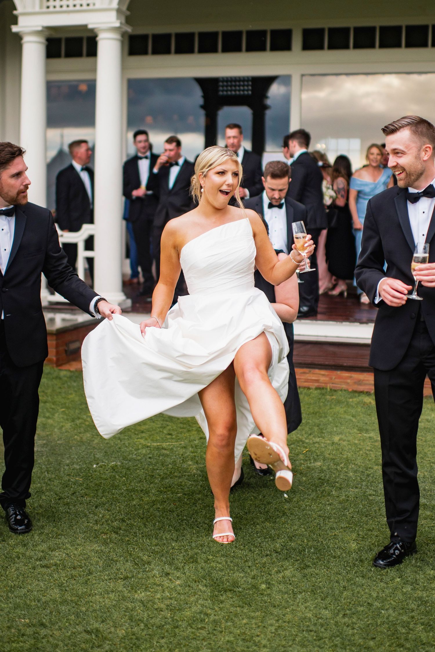 Wedding party having fun on lawn as bride kicks up her dress playfully.