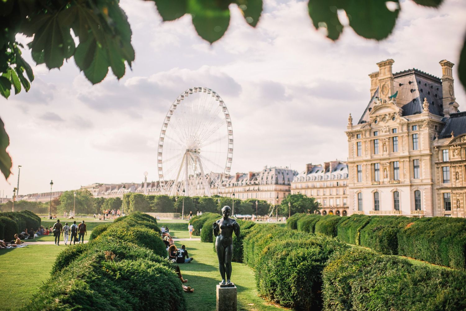 Historic buildings and a Ferris wheel visible through manicured hedges in a European city park.