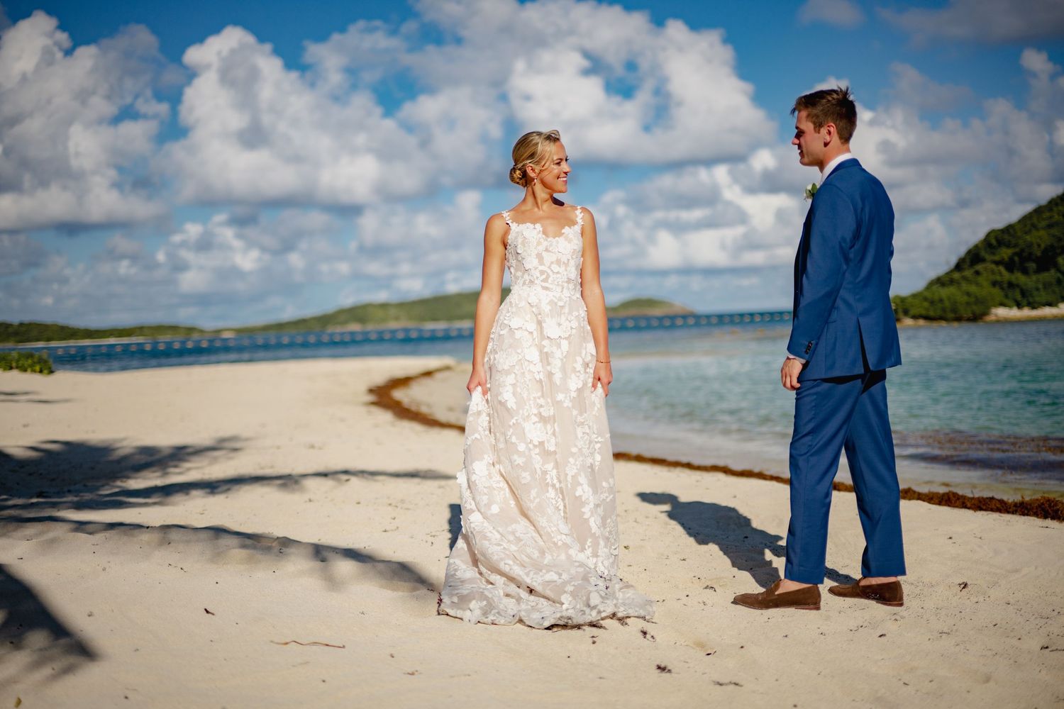 A couple in wedding attire stands on a pristine beach with turquoise water and dramatic clouds in the background.