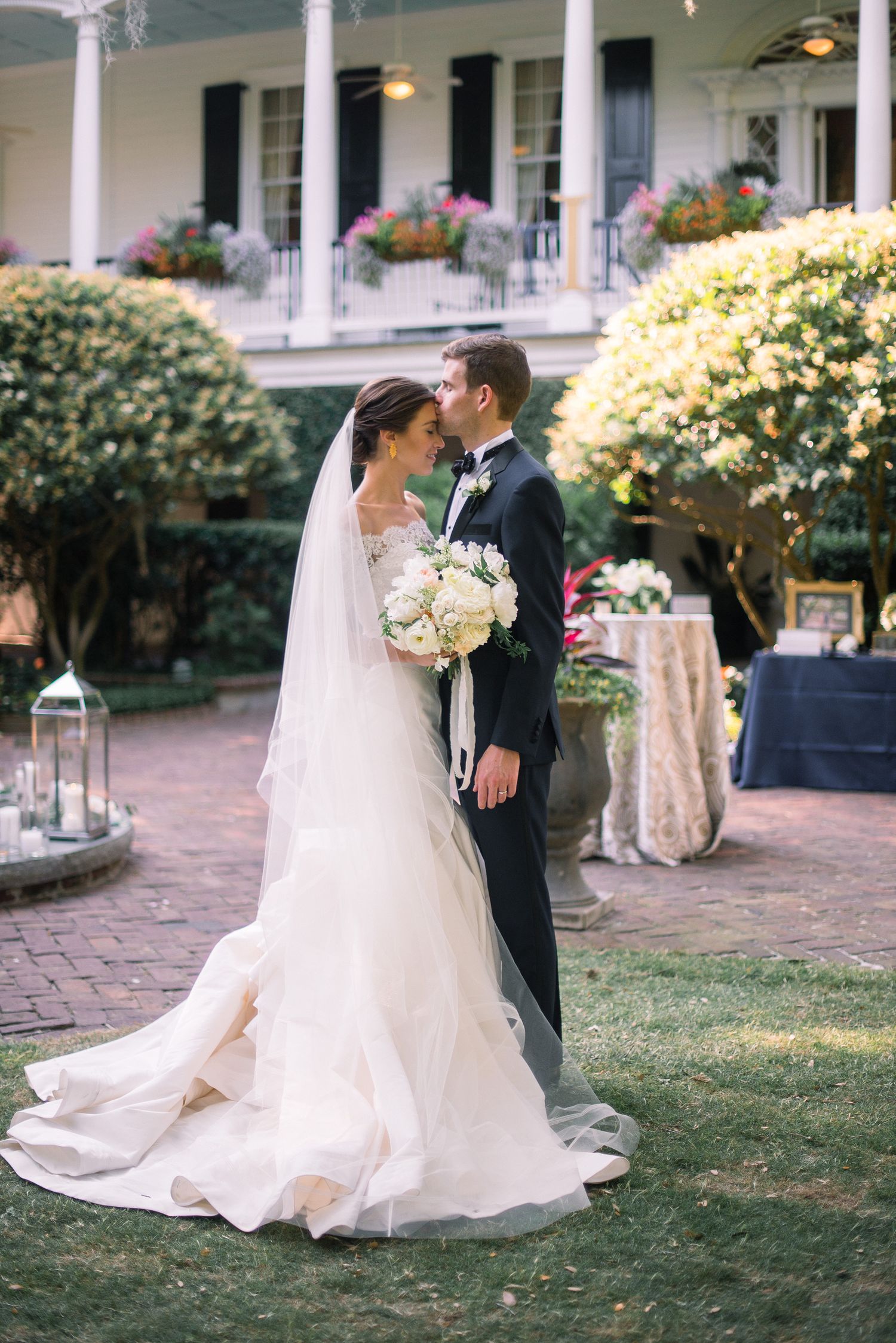 Elegant wedding couple poses in a garden setting with white columns and blooming flowers in the background.