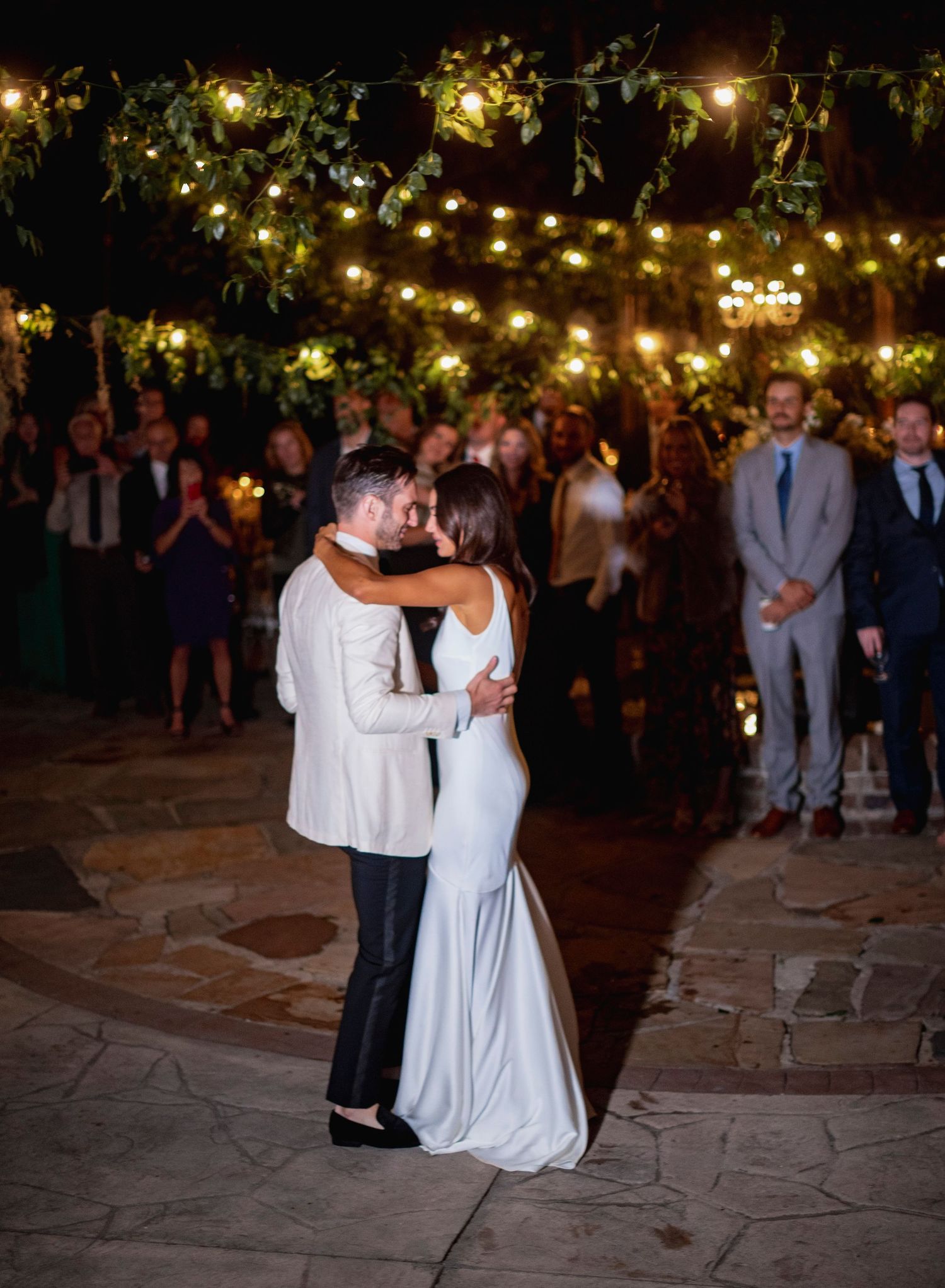 A couple shares their first dance under romantic string lights at an outdoor evening wedding reception.