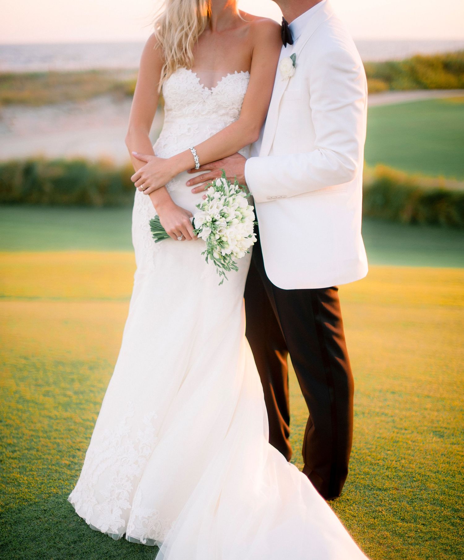 Newlywed couple embraces on a golf course at sunset while holding a delicate white floral bouquet.