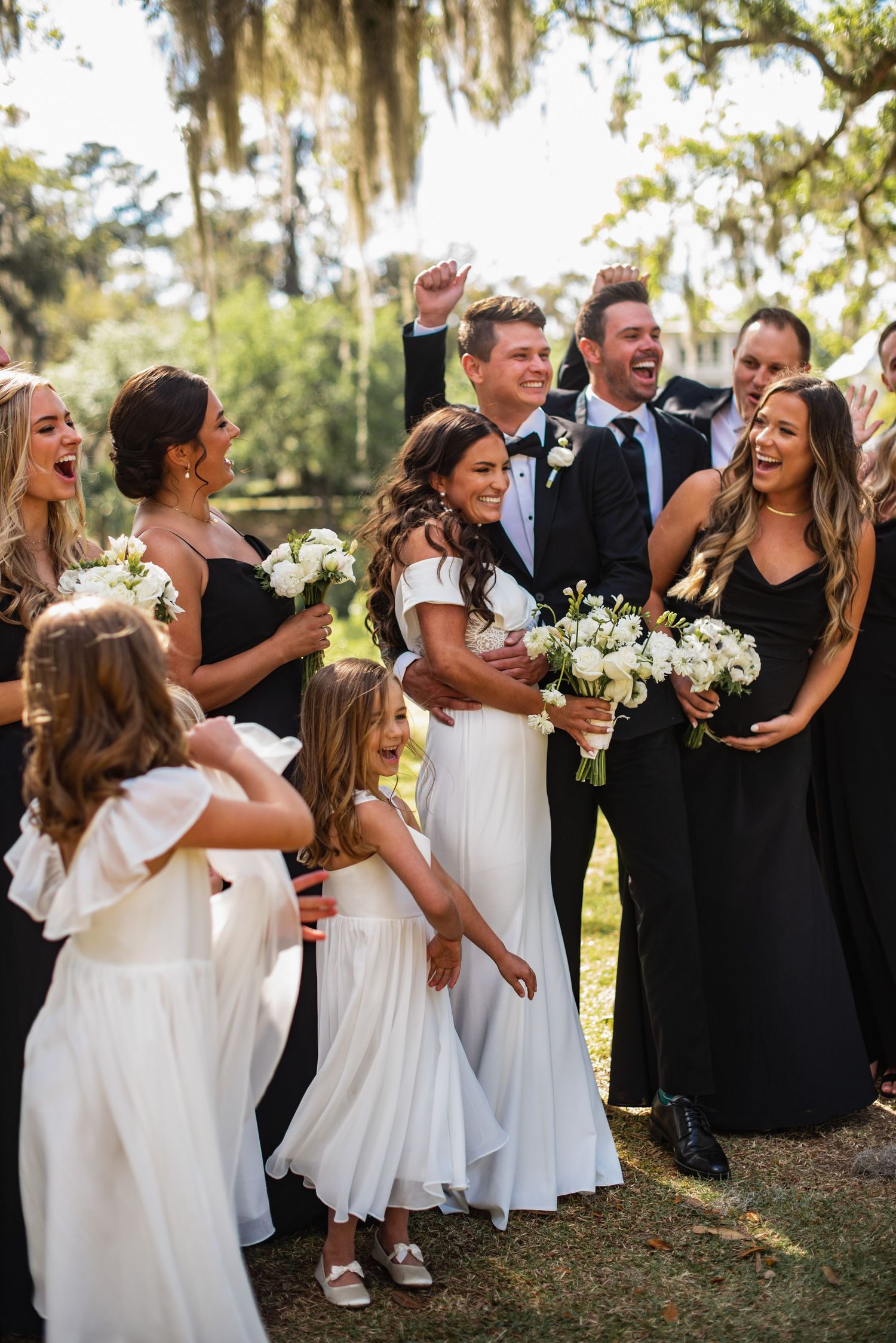 Wedding party laughs together during outdoor photo session with Spanish moss hanging from trees in background.
