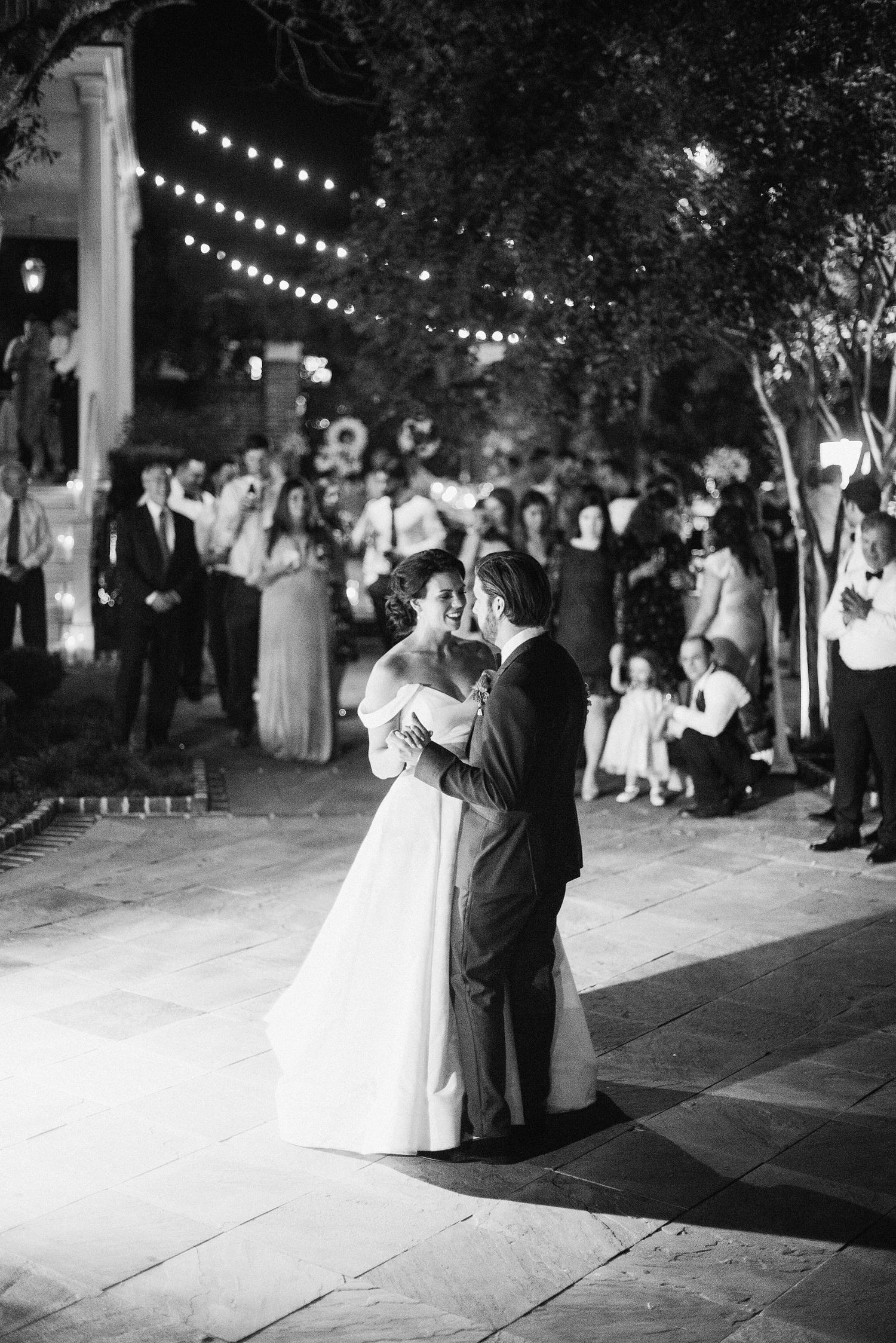 A couple shares a romantic dance under twinkling string lights at an outdoor evening wedding reception.
