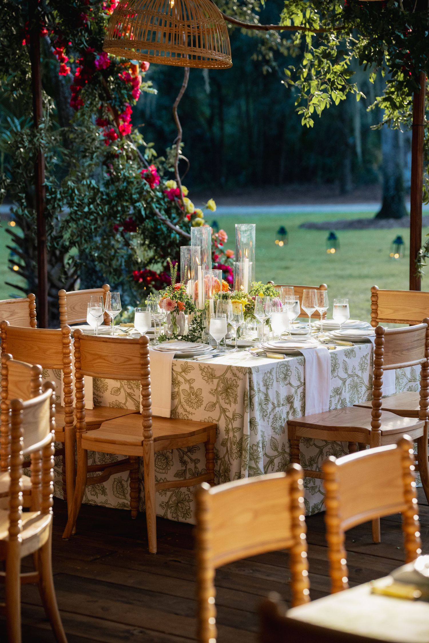 Elegant outdoor dining table with wooden chairs and floral tablecloth set under hanging lantern with climbing roses nearby.