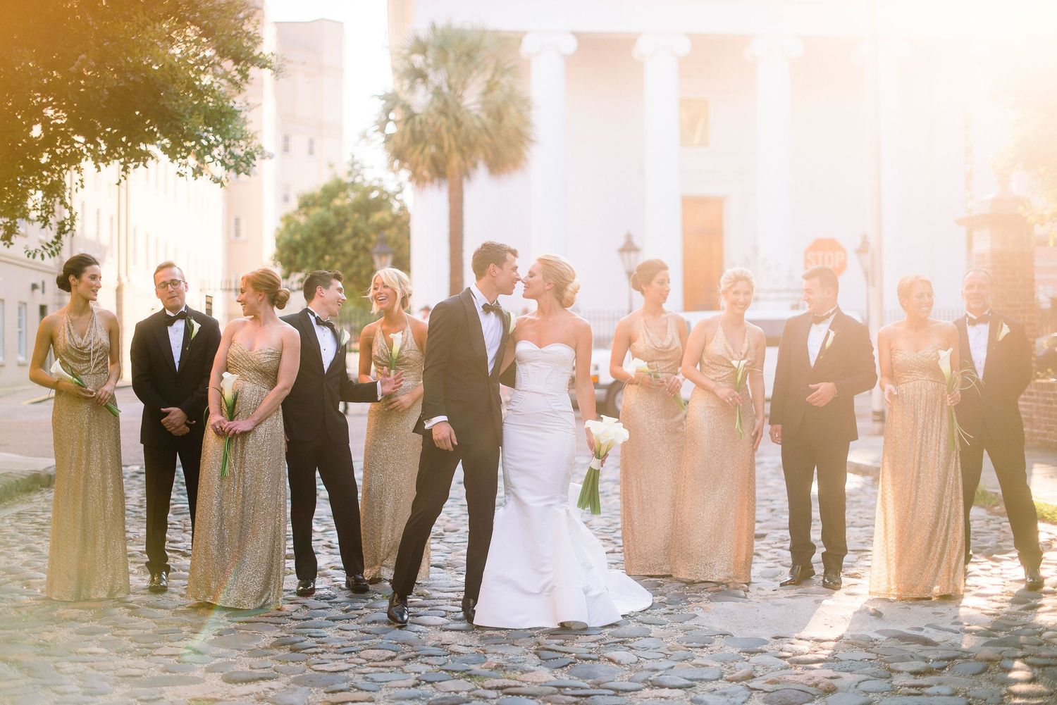 Wedding party poses together on cobblestone street during golden hour sunset with palm trees in background.