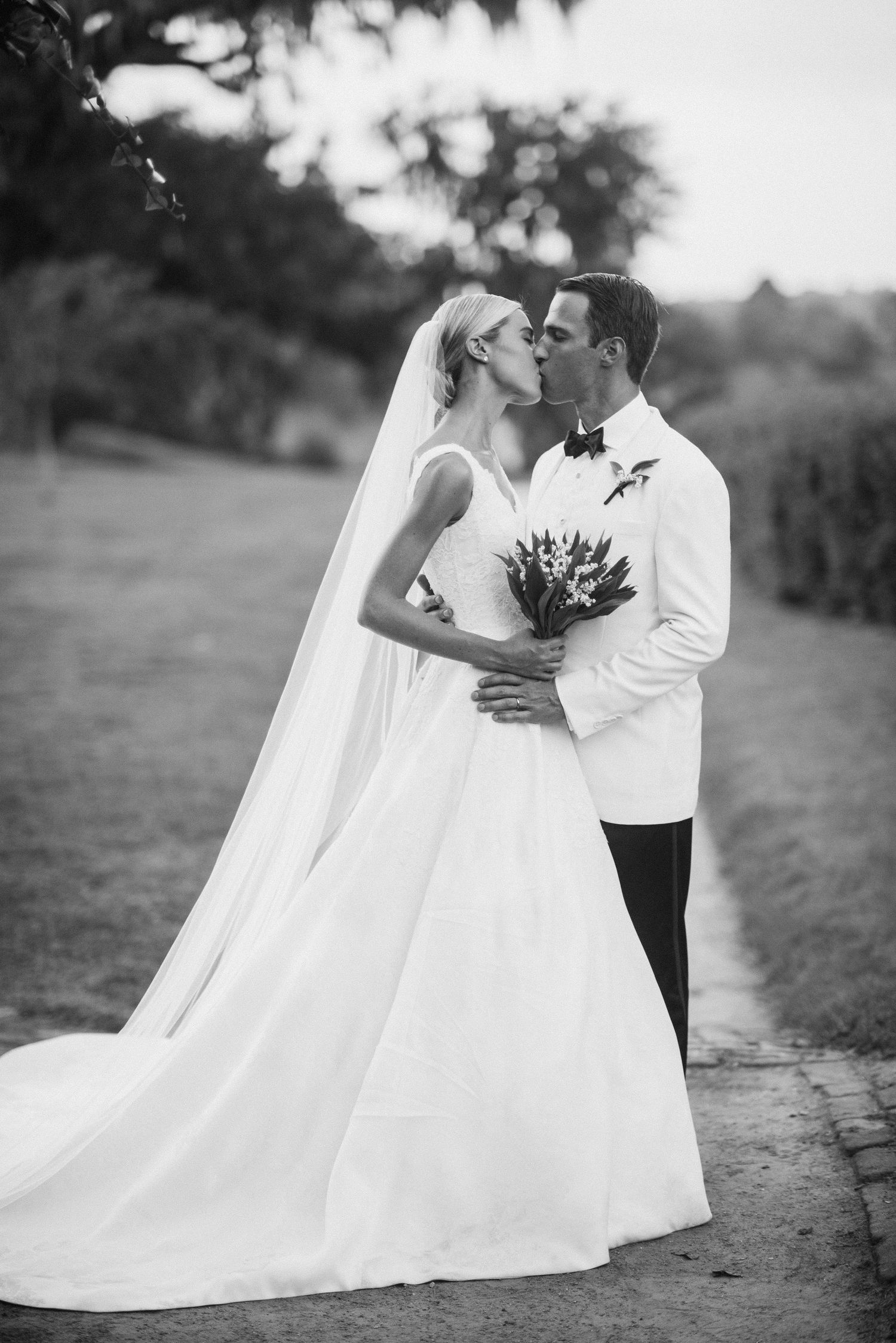 A romantic black and white wedding photo of a couple kissing outdoors while the bride holds a bouquet.