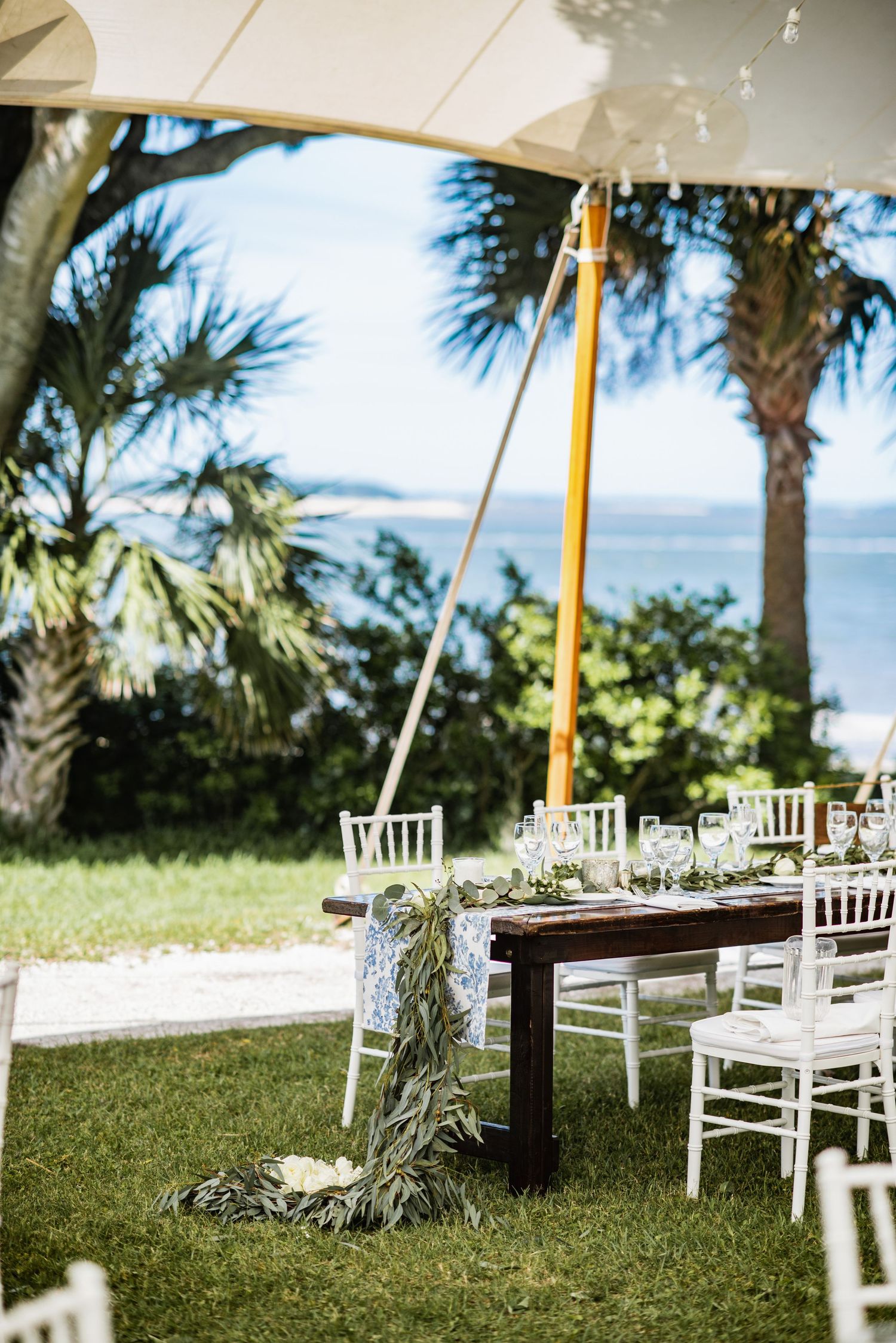 White wooden chairs arranged on a grassy lawn for an outdoor wedding ceremony with palm trees and ocean views.