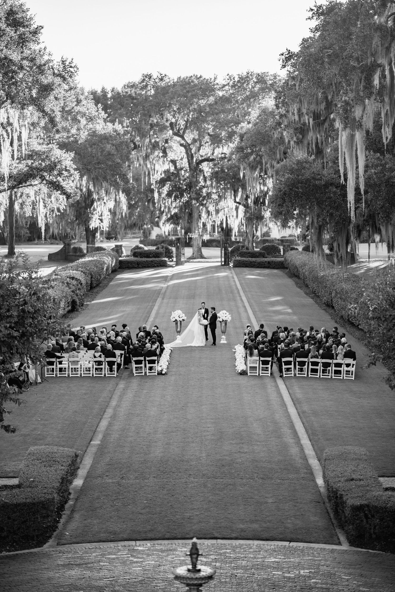 A black and white photo shows an outdoor wedding ceremony setup with rows of chairs on a manicured lawn beneath oak trees.