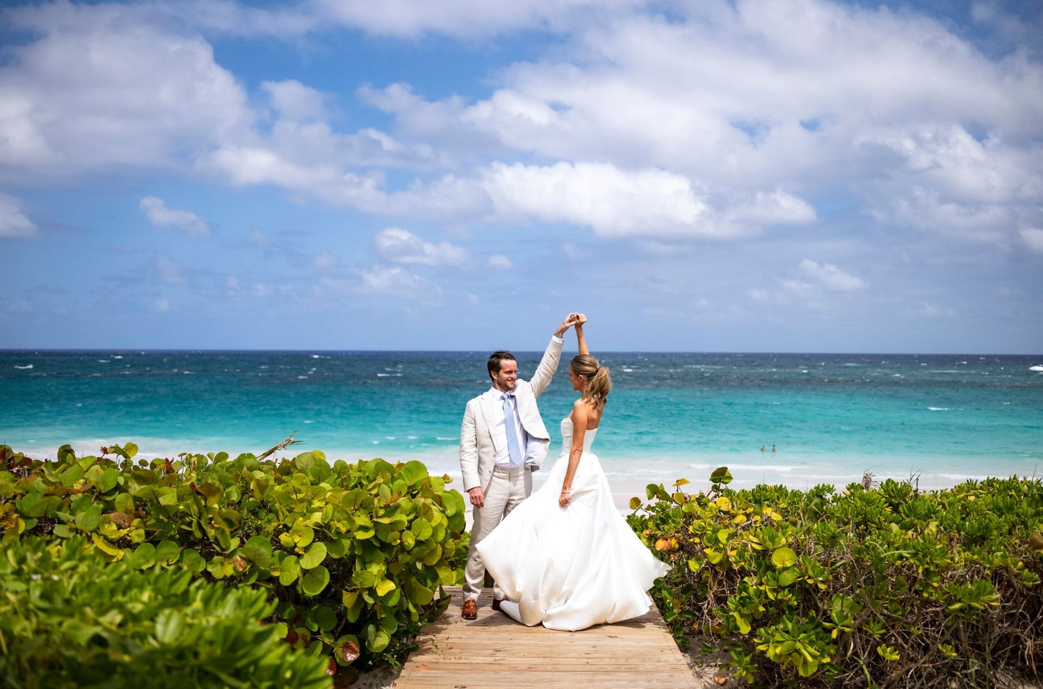 Newlyweds pose on a coastal pathway with turquoise ocean waters of harbour island Bahamas and tropical foliage in the background.