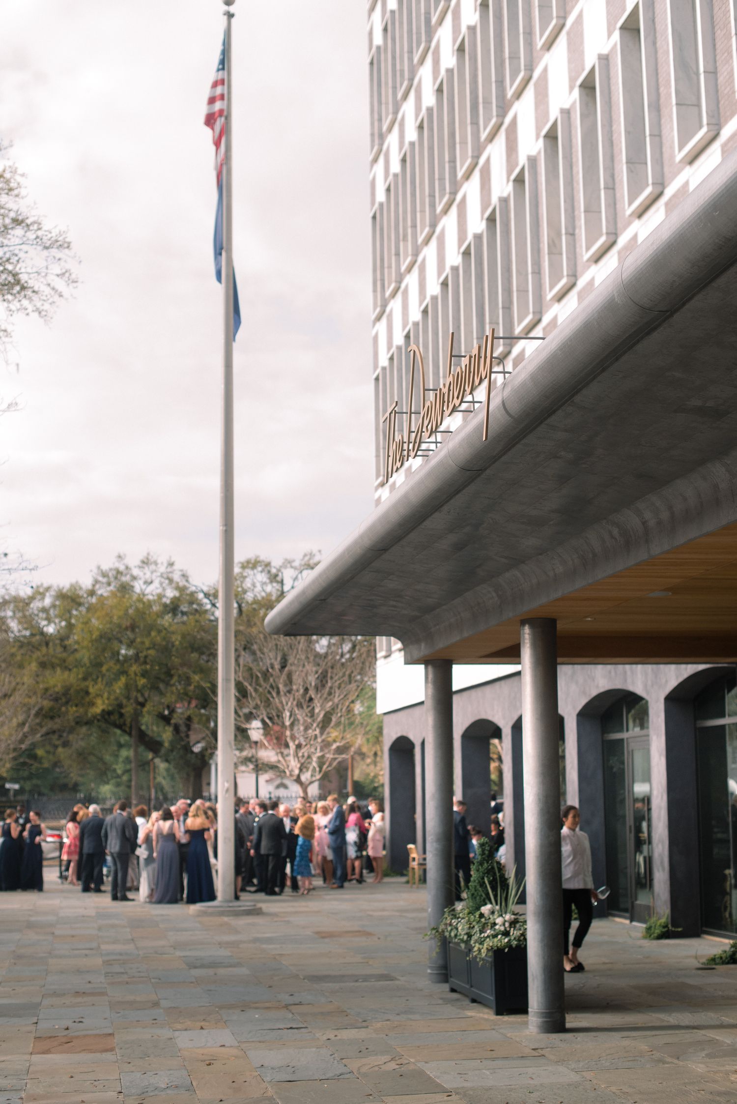 People wait in line outside a modern government building with columns and American flags flying overhead.