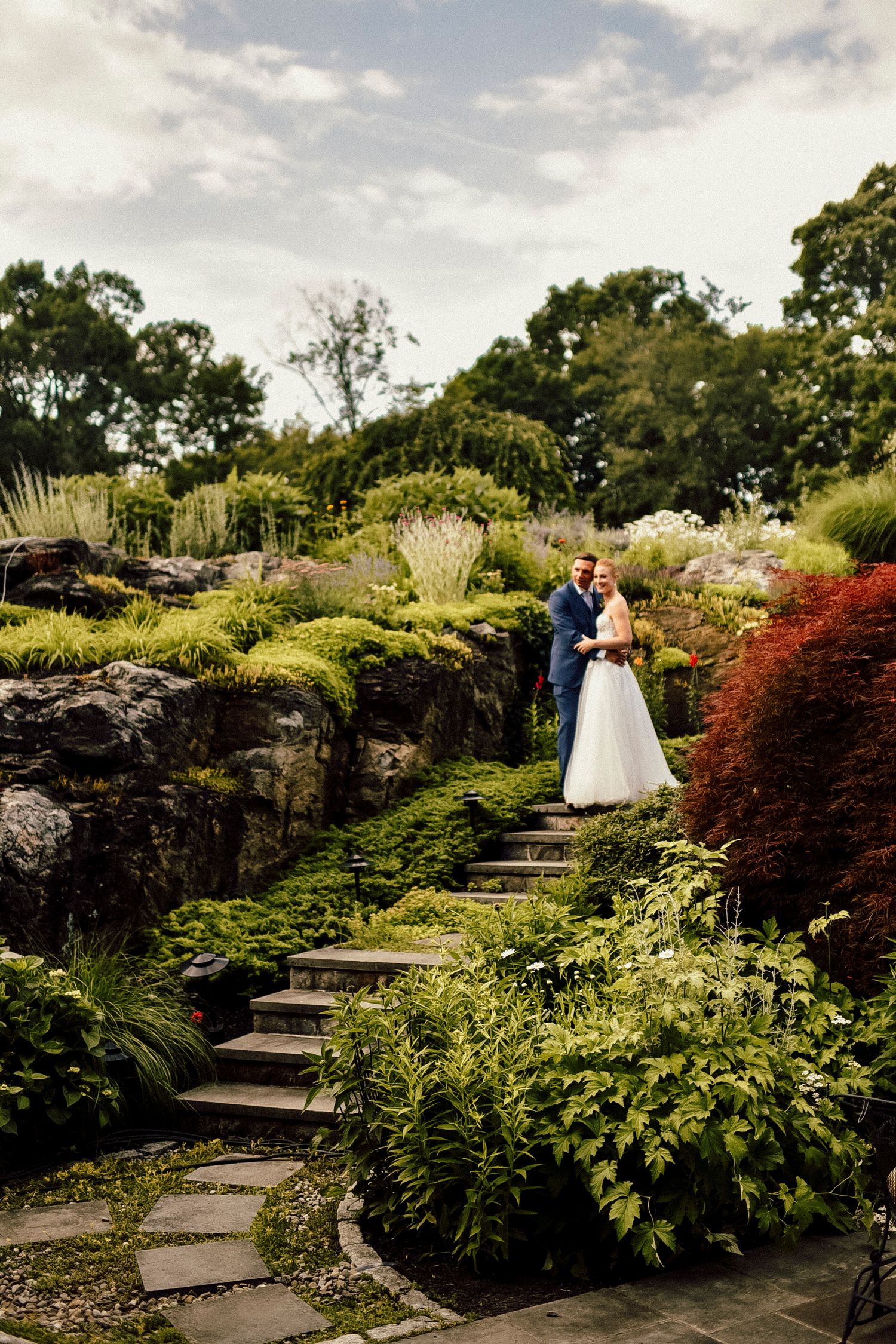 A series of stone steps wind through a lush garden with red maples and flowering shrubs on a cloudy day.