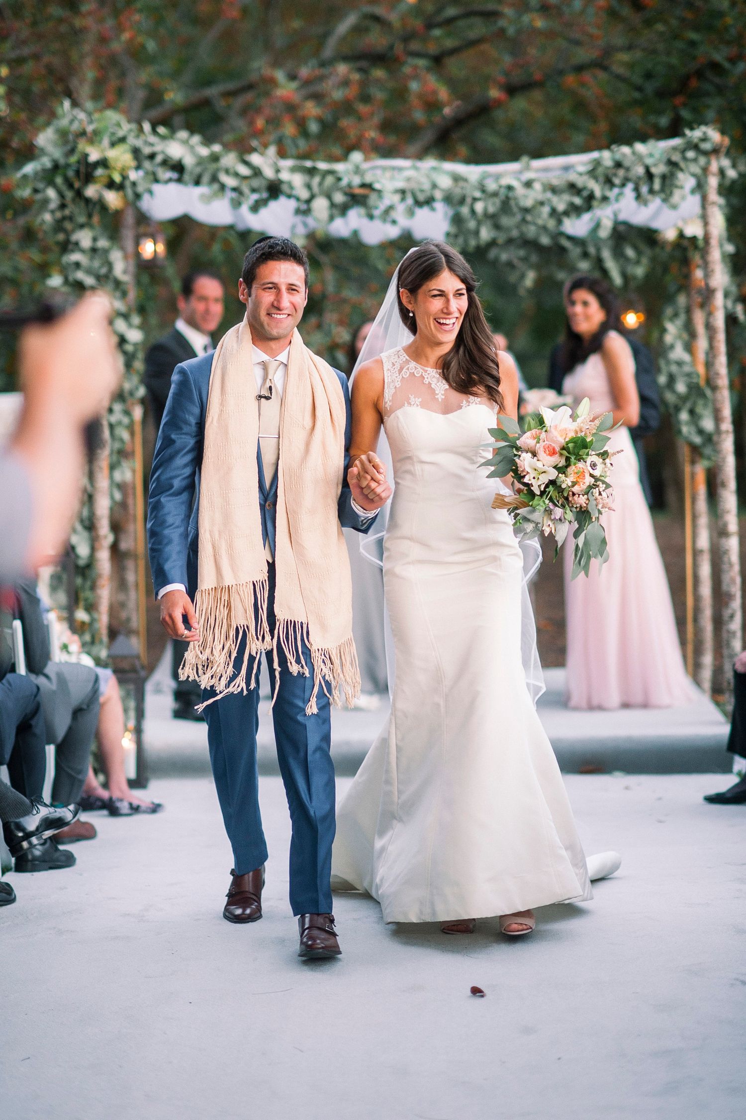 Wedding party members walking and celebrating outdoors under string lights and trees.