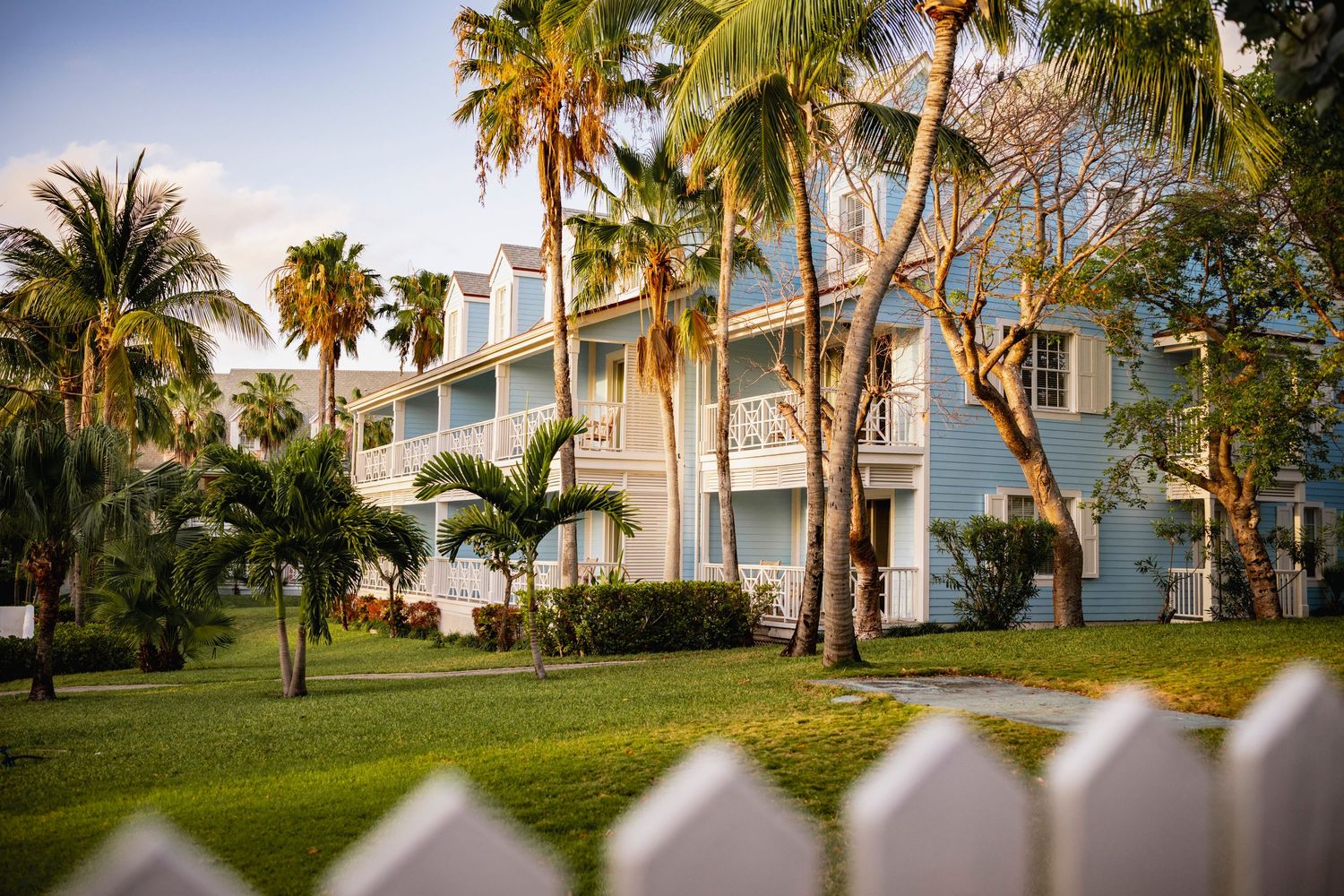 Tropical resort building in Harbour island with white architecture and palm trees viewed through a white picket fence.