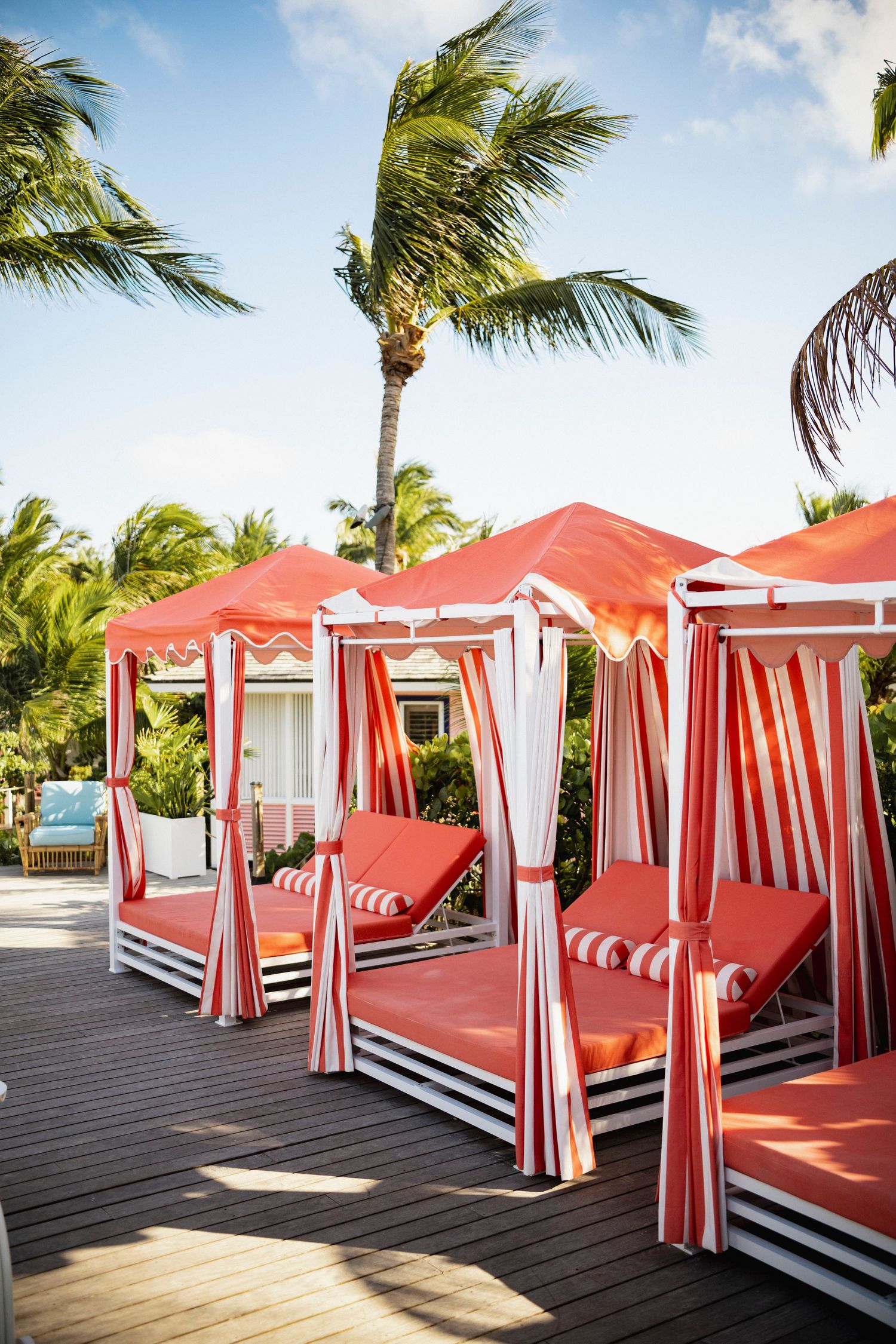 Luxurious red and white striped cabanas with daybeds line a wooden deck under swaying palm trees at a Coral Sands resort.