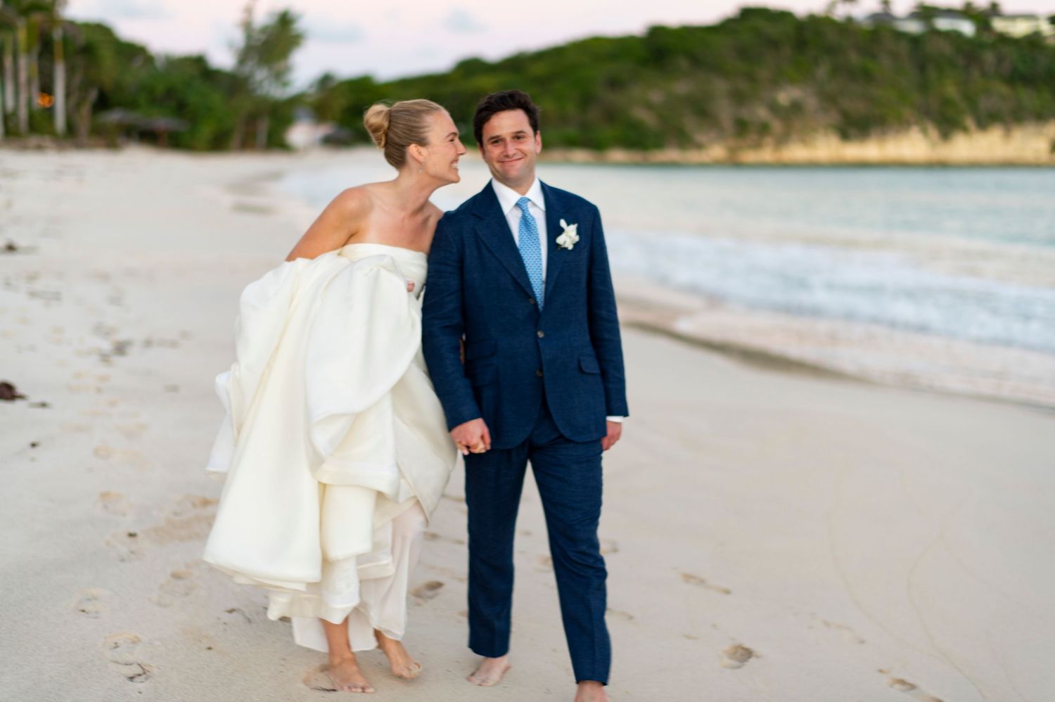 A couple walks along a scenic beach shoreline during sunset wearing formal wedding attire.