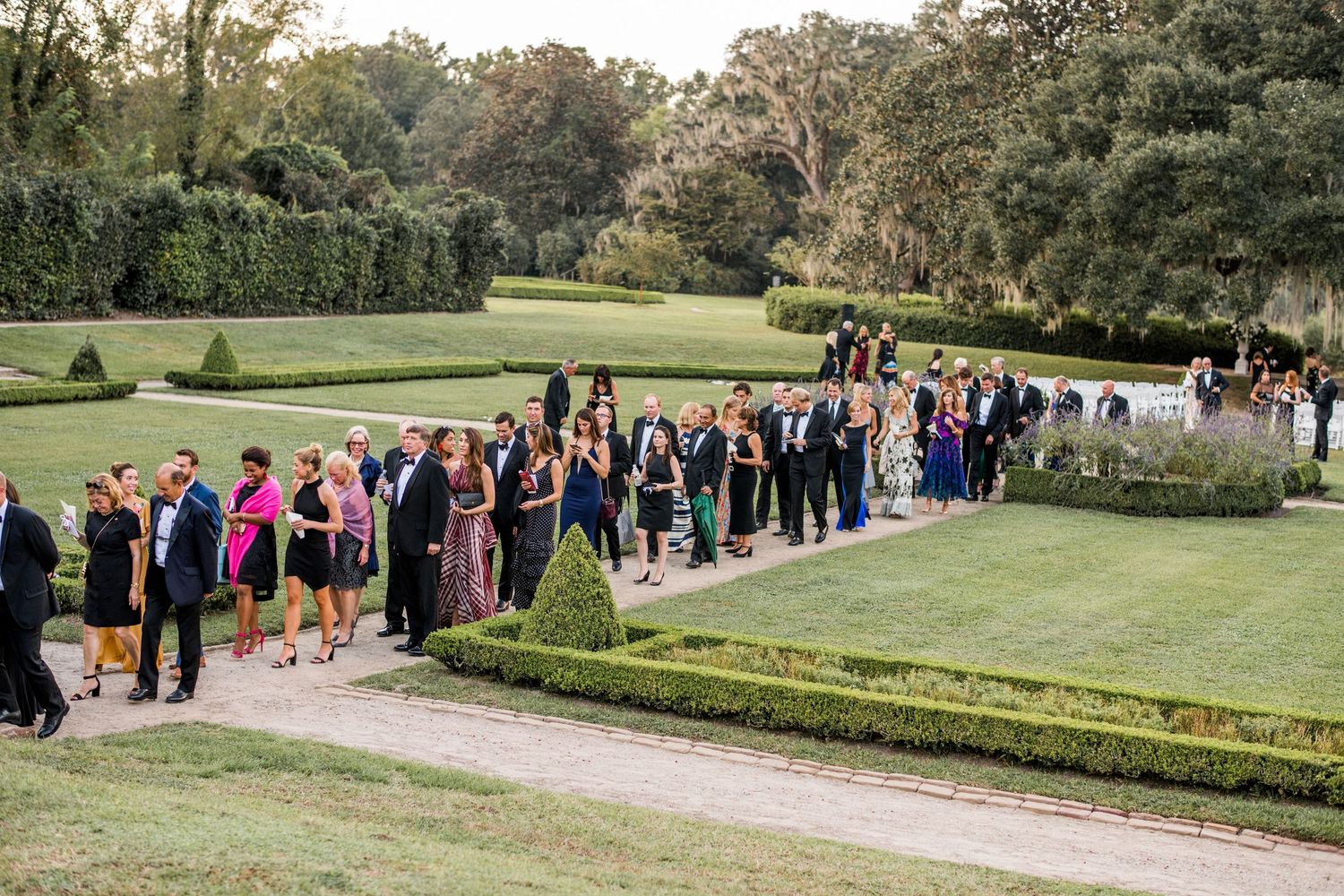 Wedding guests line up along a scenic garden path during an elegant outdoor ceremony at a manicured estate.
