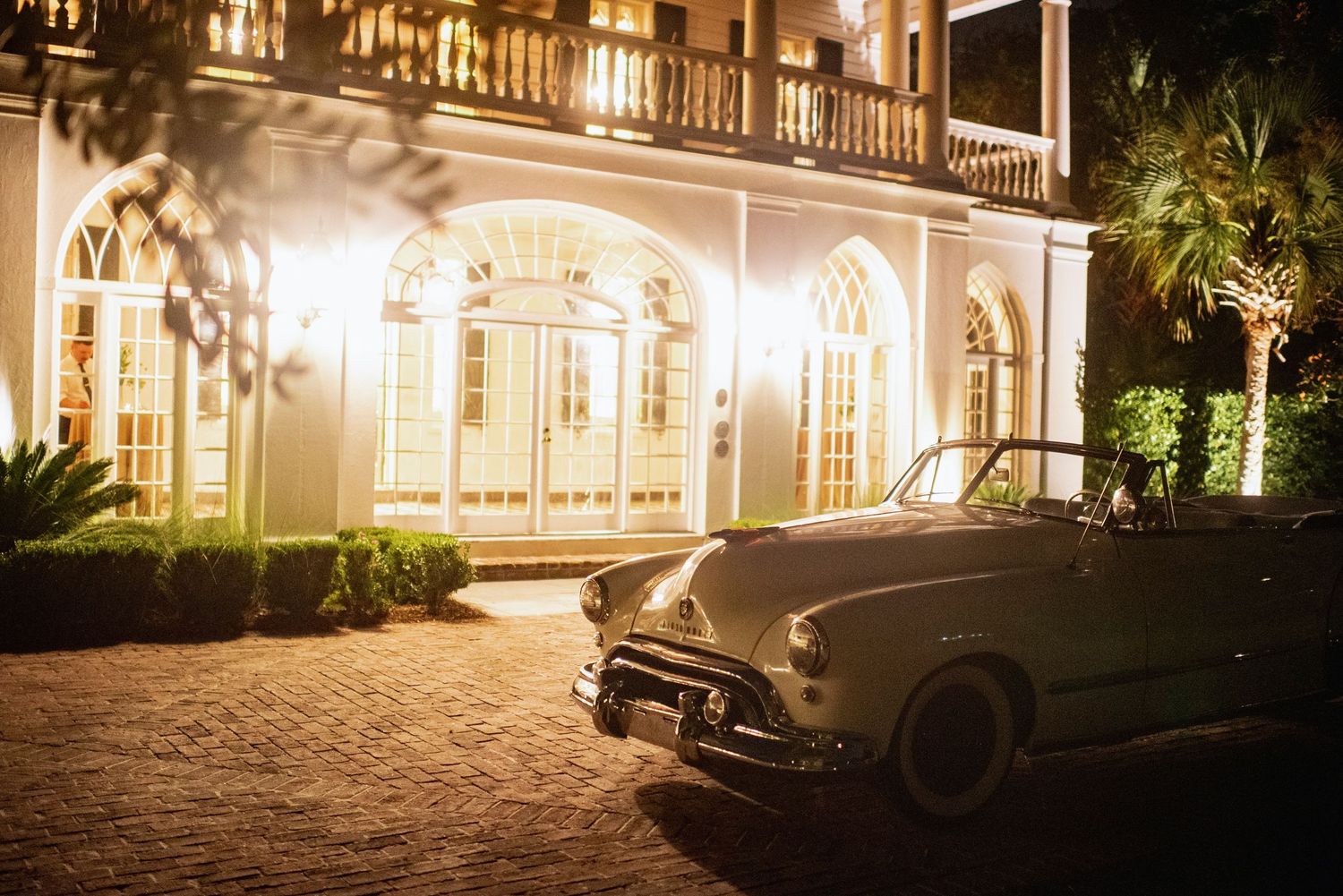 Vintage car parked outside illuminated southern mansion with ornate windows and balcony at night.