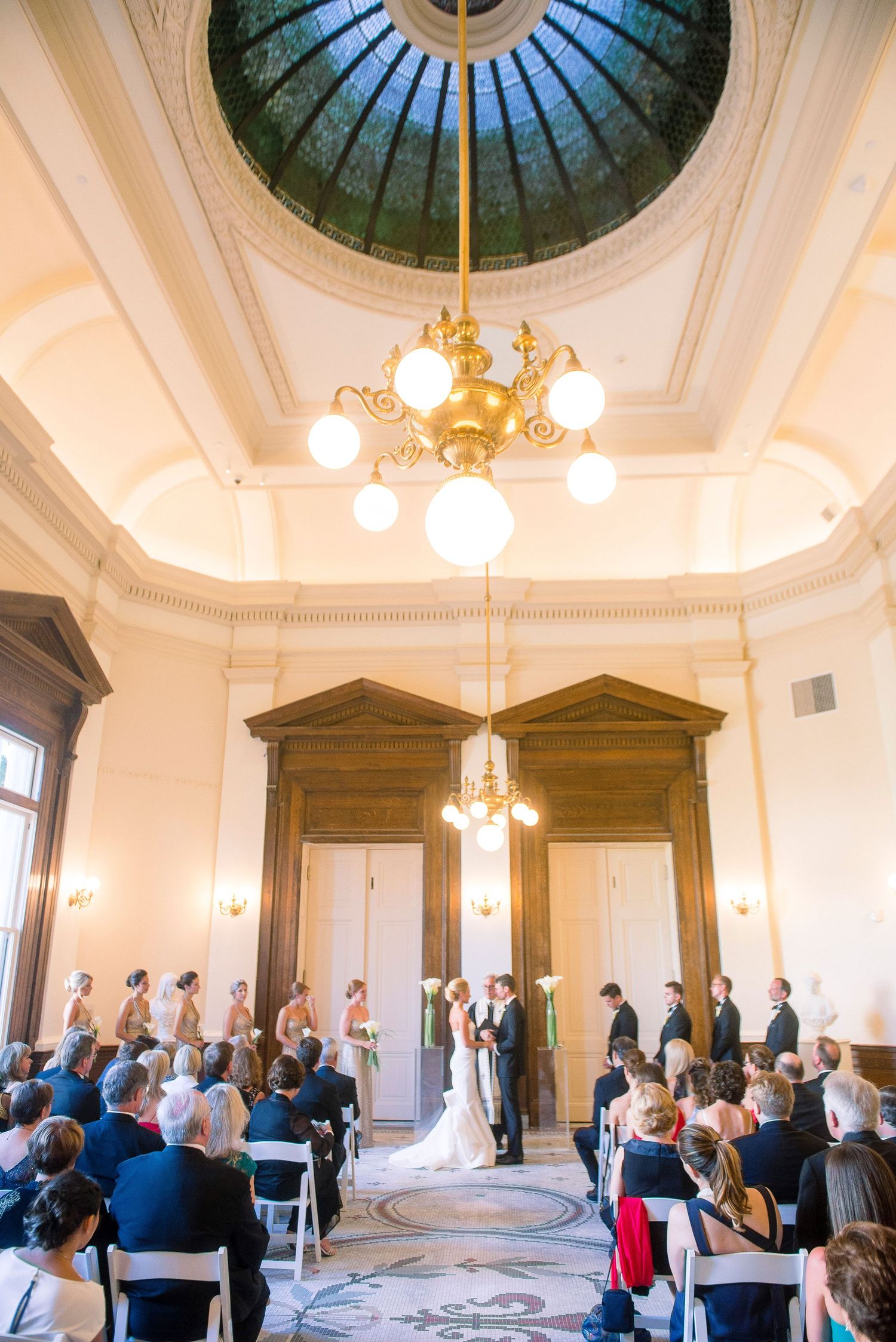 Elegant wedding ceremony in a historic domed room with ornate architectural details and chandeliers.