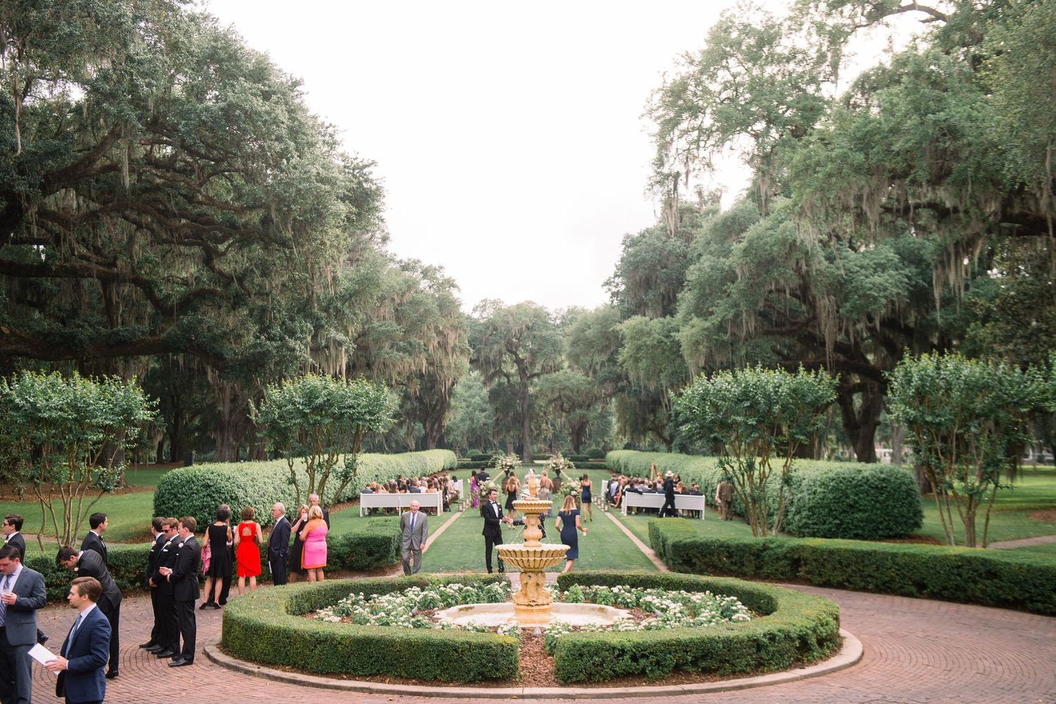 A panoramic view of a formal garden path lined with oak trees and manicured hedges during an outdoor event.