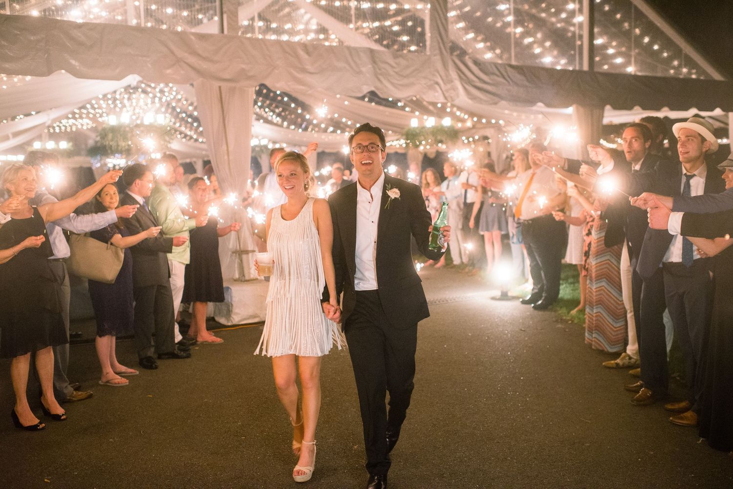 Wedding guests form a sparkler tunnel exit under a white tent as newlyweds walk through during evening reception celebration.