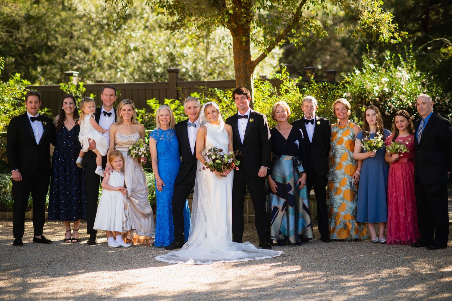Wedding party and guests gather outdoors for group photos in a sunlit garden setting.