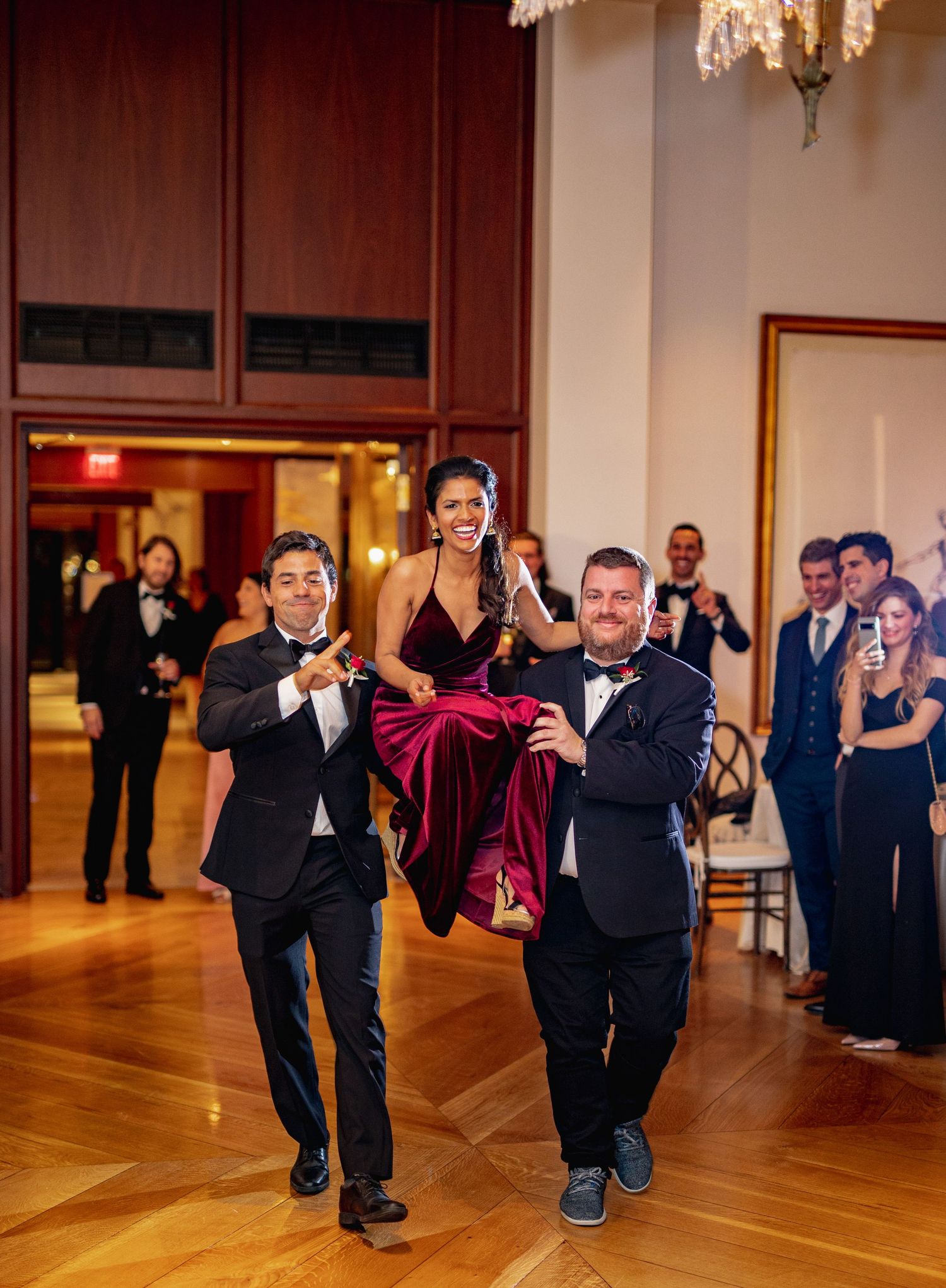 Groomsmen perform synchronized dance moves in a ballroom with wooden floors and elegant chandeliers.
