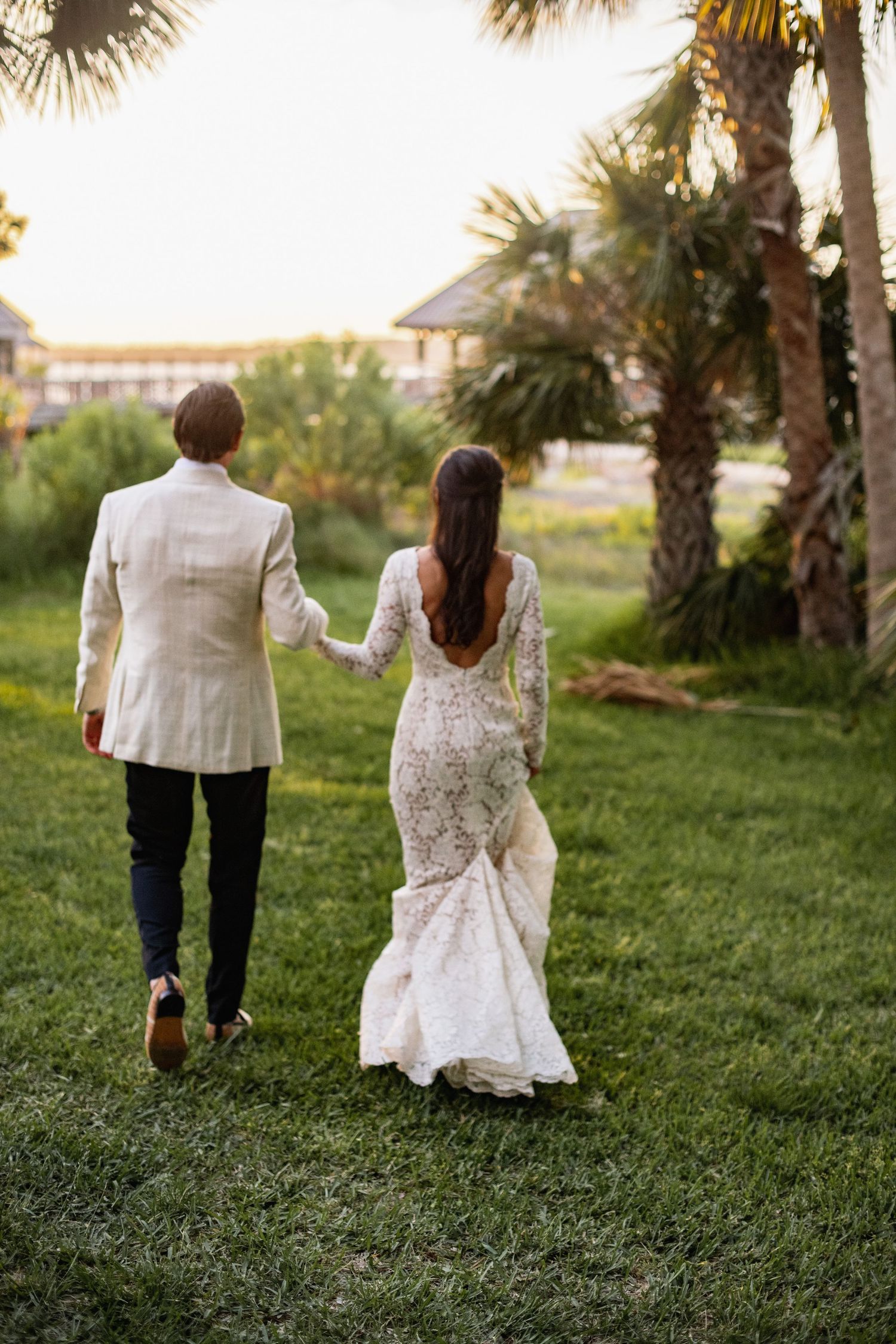 A couple walks hand in hand through a lush green lawn with palm trees in the background at sunset.