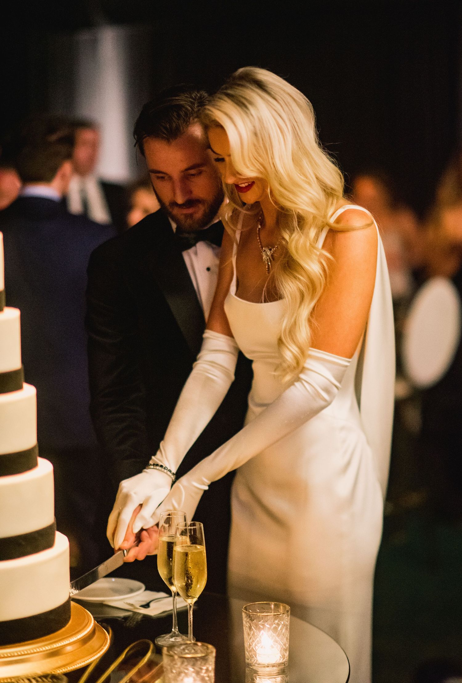 A couple in formal attire cuts into an elegant black and white tiered wedding cake at a candlelit reception.