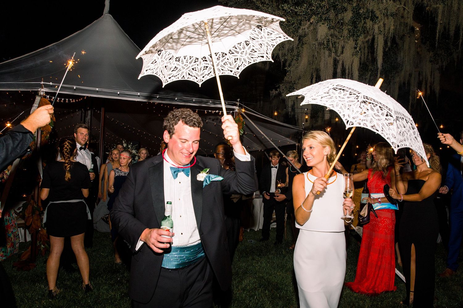 Wedding guests dance and celebrate under white lace parasols at an outdoor evening reception.