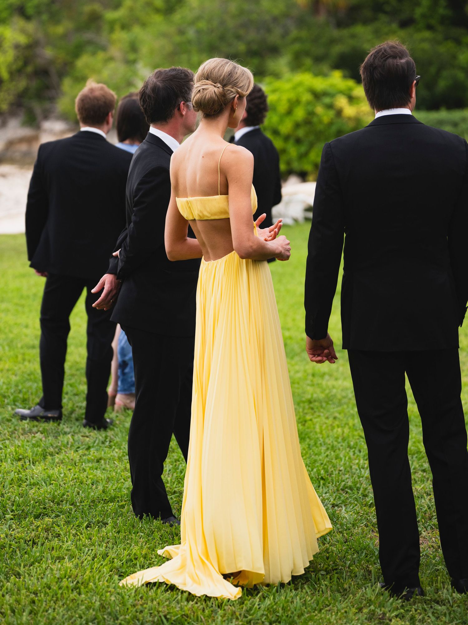 Wedding party members in black suits and yellow dresses gather on green lawn during outdoor ceremony.