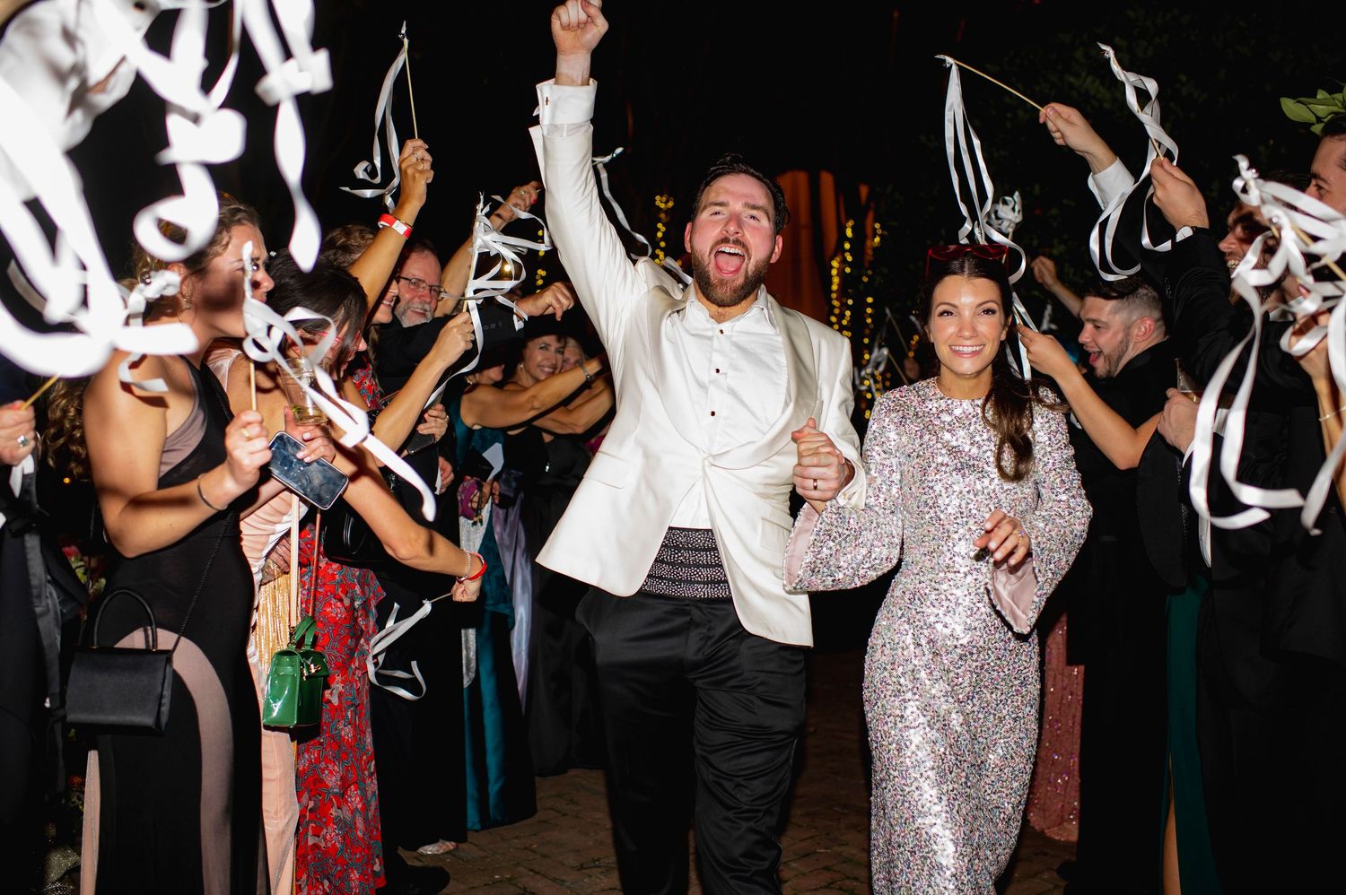 A group of wedding guests dance and celebrate during a reception with sparklers and lights in the background.