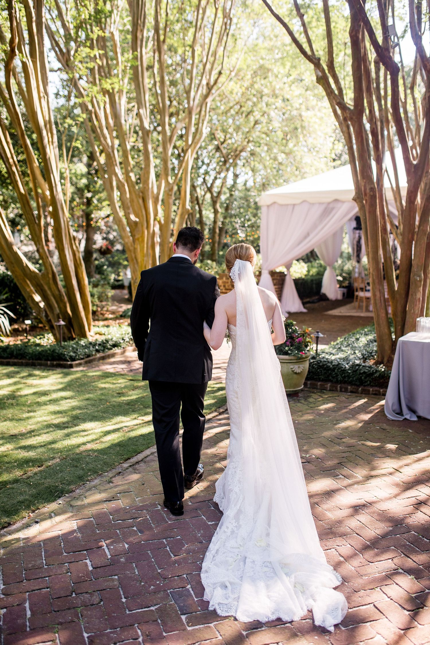 Wedding couple walking along a brick path under a canopy of trees in a garden setting.