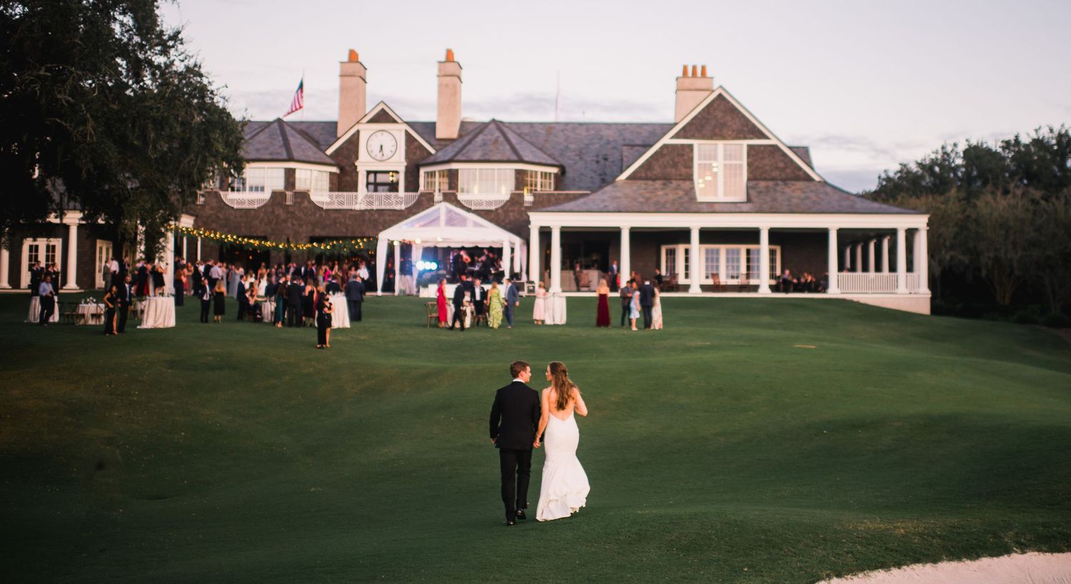 Elegant wedding venue panorama showing a grand clubhouse with white columns and expansive green lawn at sunset.